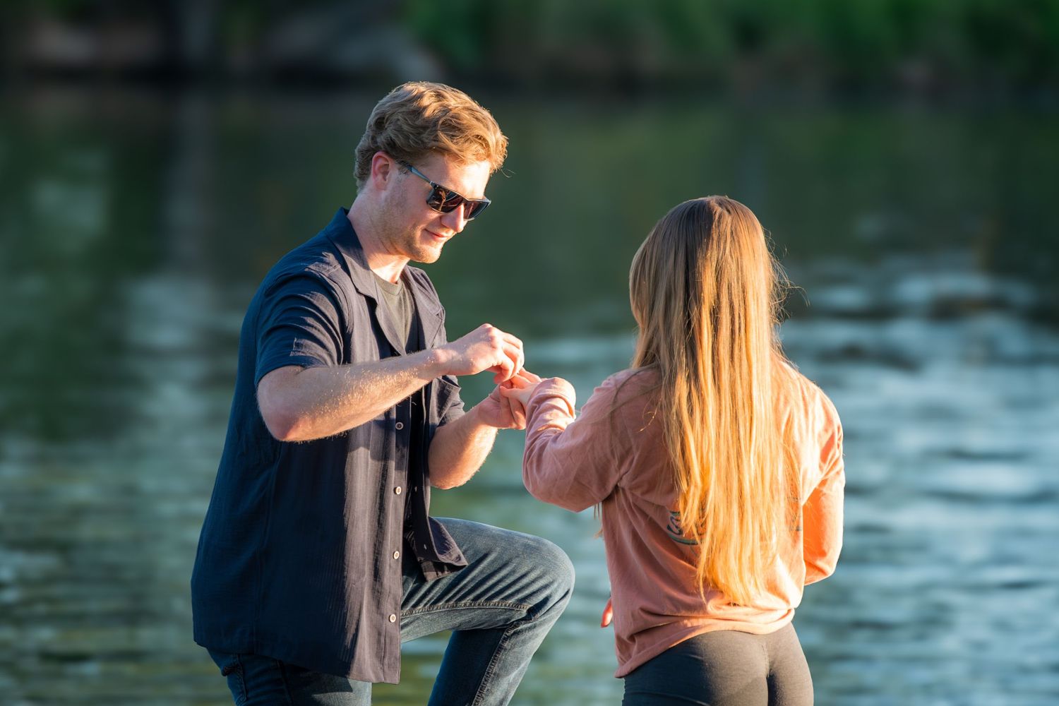 Man placing engagement ring on womans finger near a scenic lakeside at sunset.