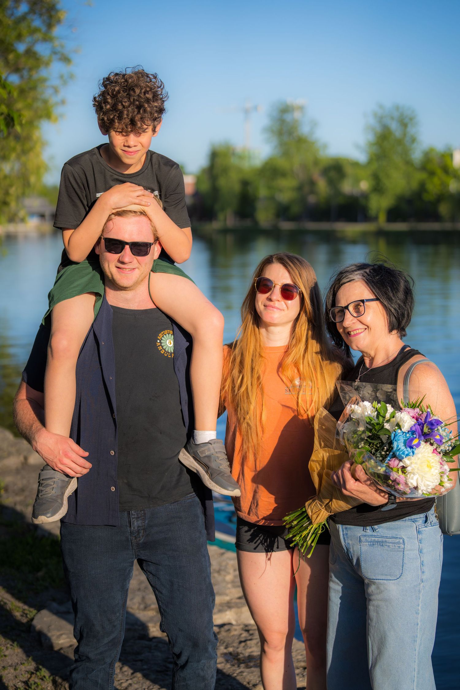 Happy family posing by a lake, man with boy on shoulders, woman holding a colorful bouquet.