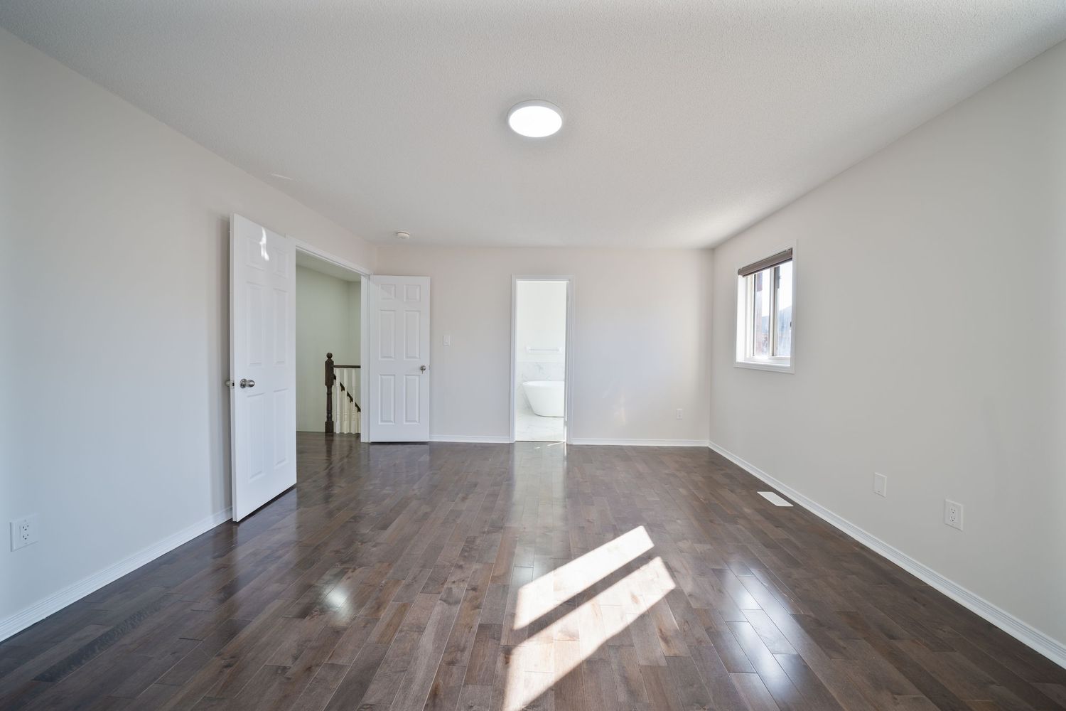Interior hallway with white walls and light hardwood flooring illuminated by recessed ceiling lights and natural sunlight.