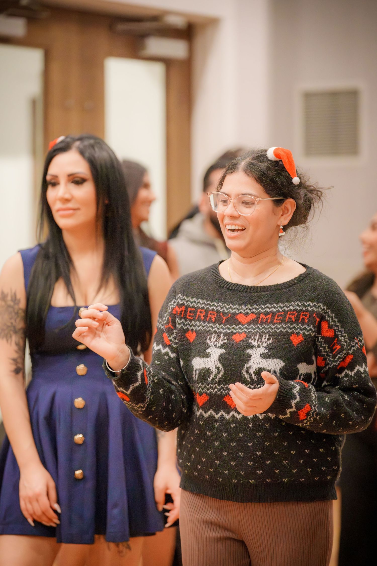 Group wearing festive holiday sweaters and outfits gather at indoor celebration.