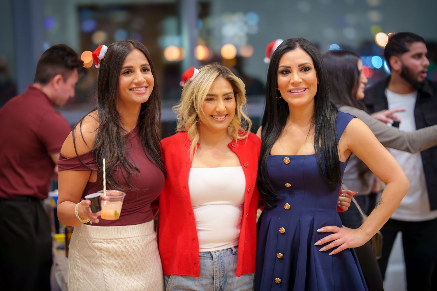Three friends pose together at a social gathering with festive drinks and attire.
