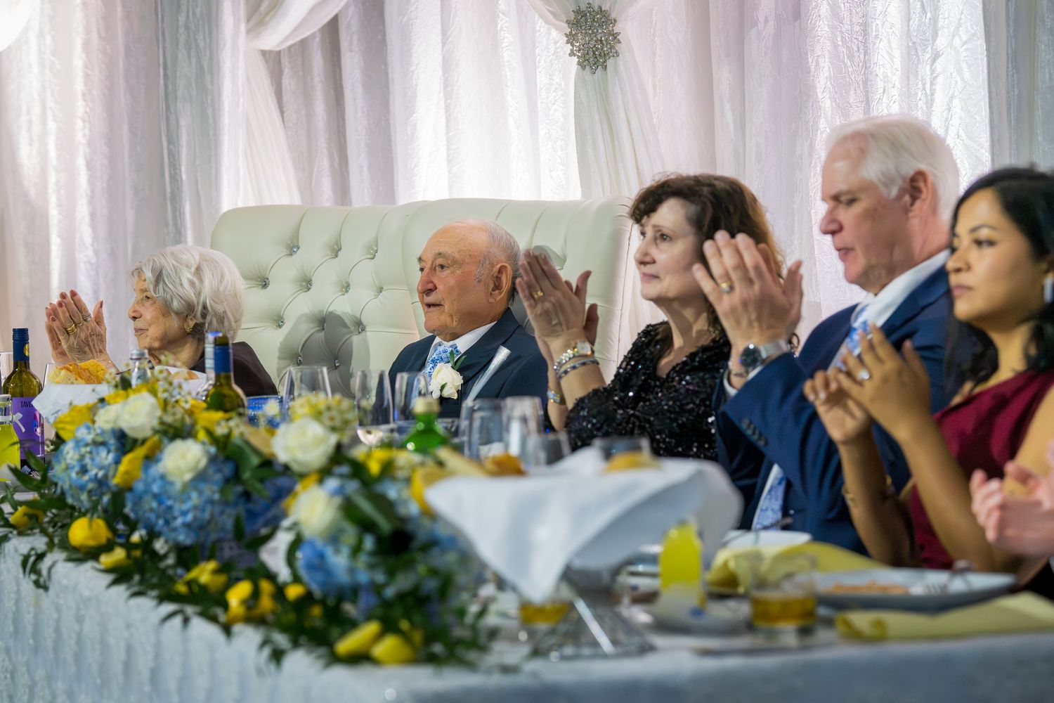 Guests applauding at a wedding reception head table adorned with blue and yellow floral arrangements.