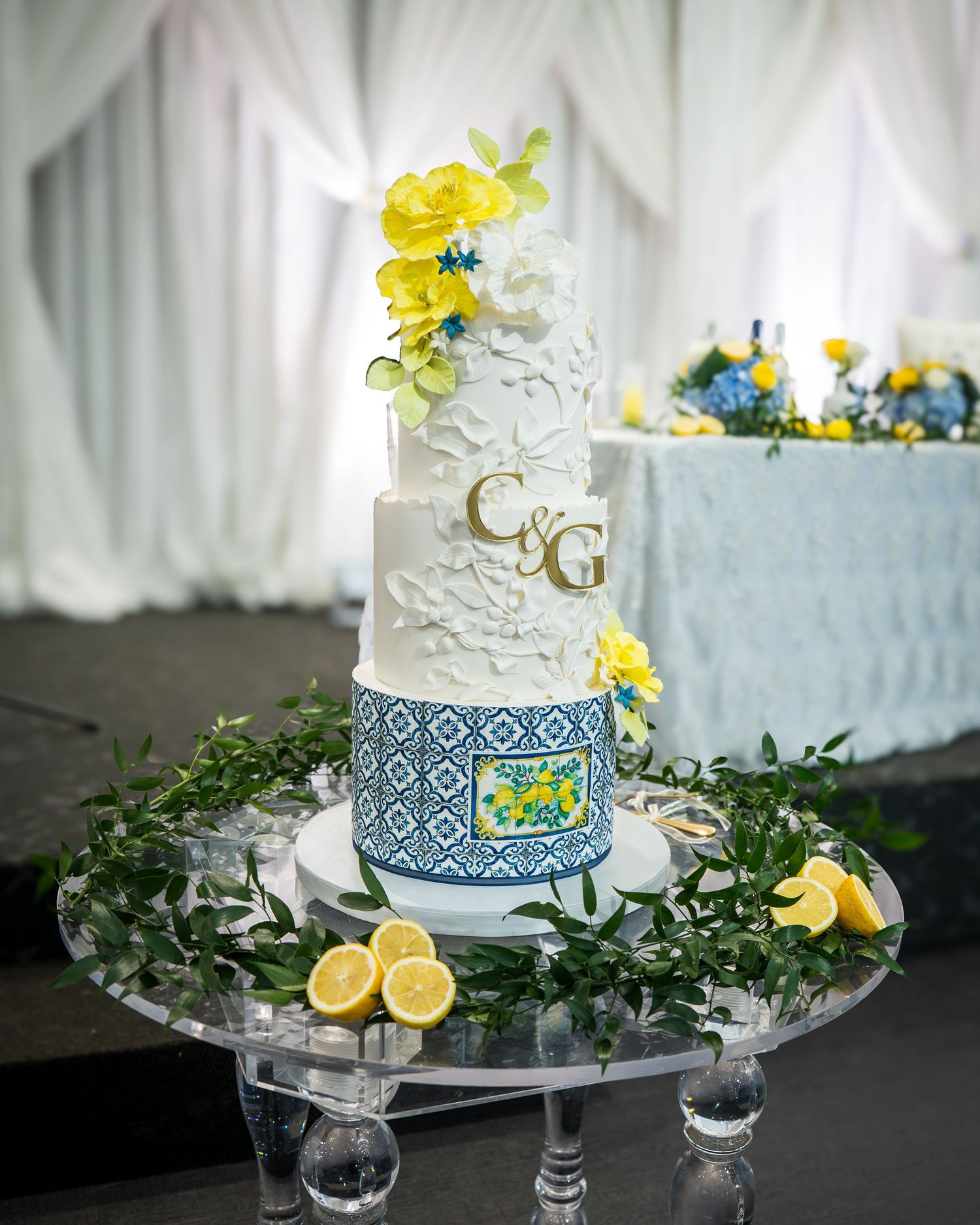 Elegant three-tier wedding cake with blue Amalfi tile design, yellow flowers, and lemon decor on a mirrored stand.