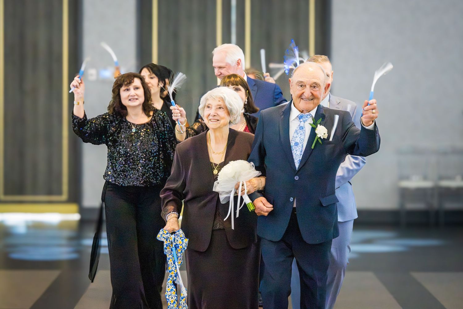 Elderly couple walking hand-in-hand with guests waving light wands at a festive anniversary celebration.