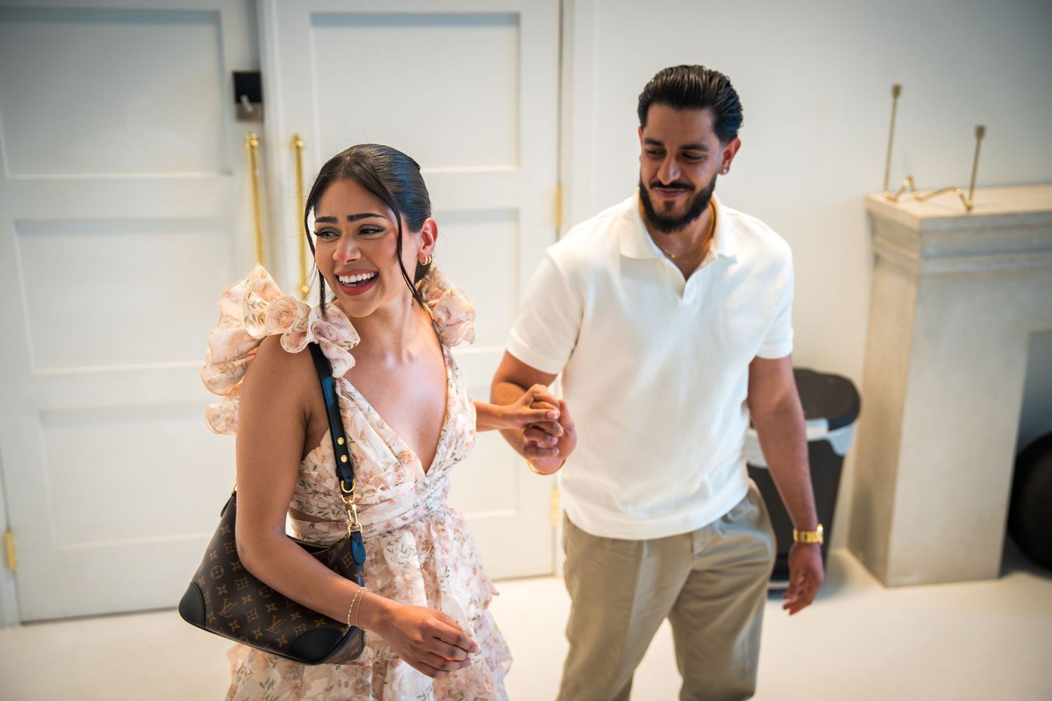 Smiling couple walking together indoors, woman in floral dress carrying designer bag, man in white polo shirt.