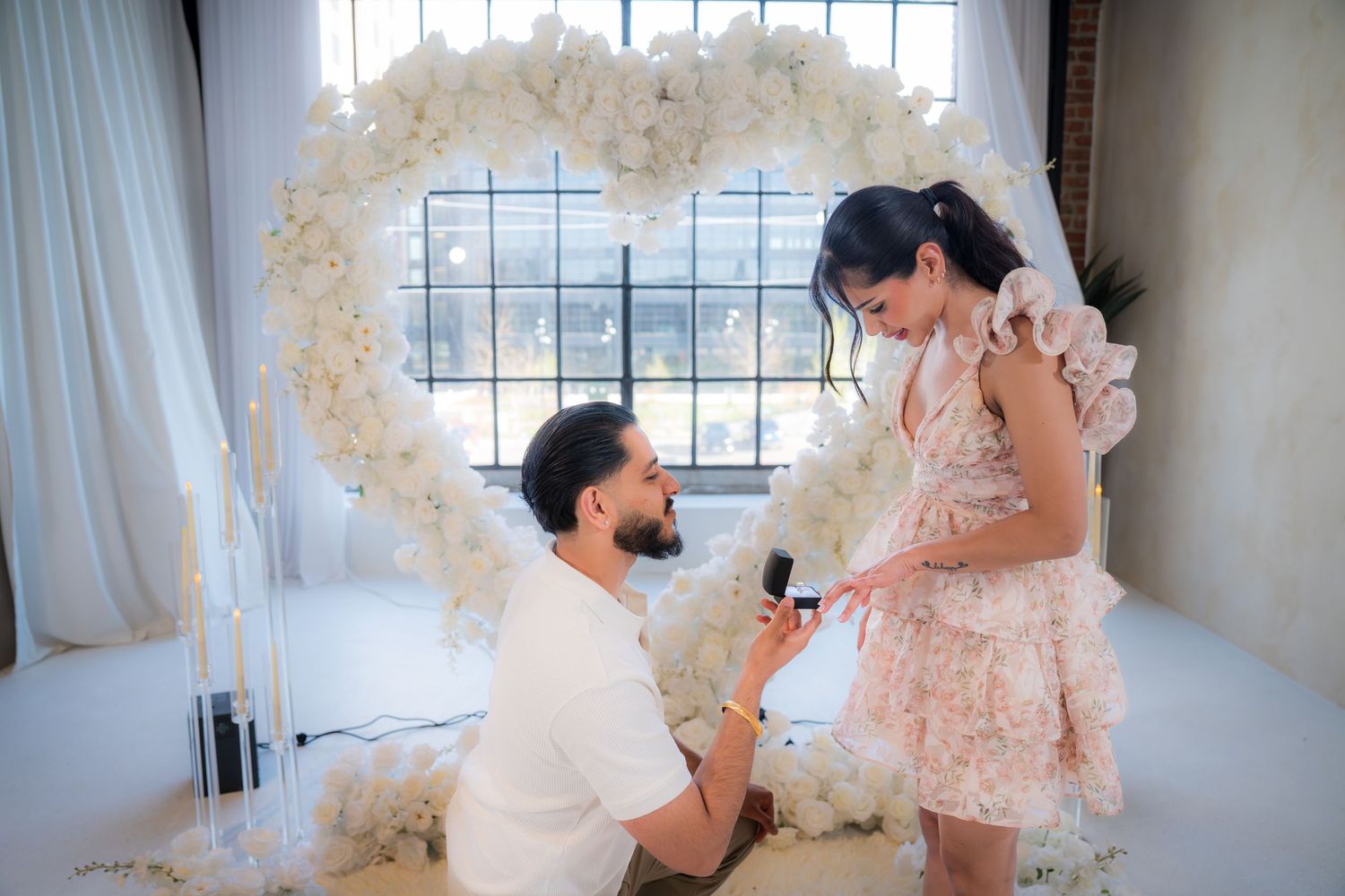 Man proposing on one knee to woman in pink dress before heart-shaped white floral arch.