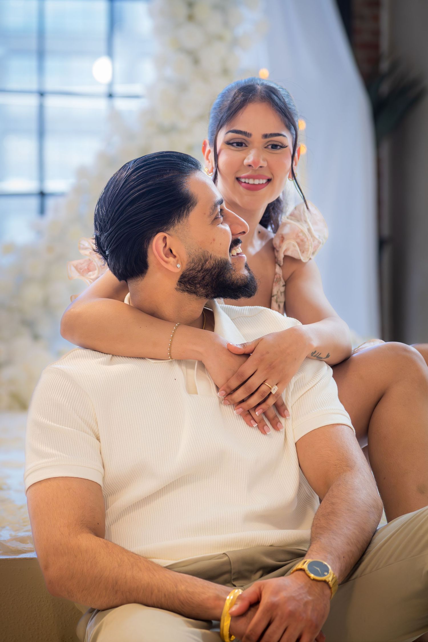 Engaged couple sitting together smiling with white floral decorations in background.