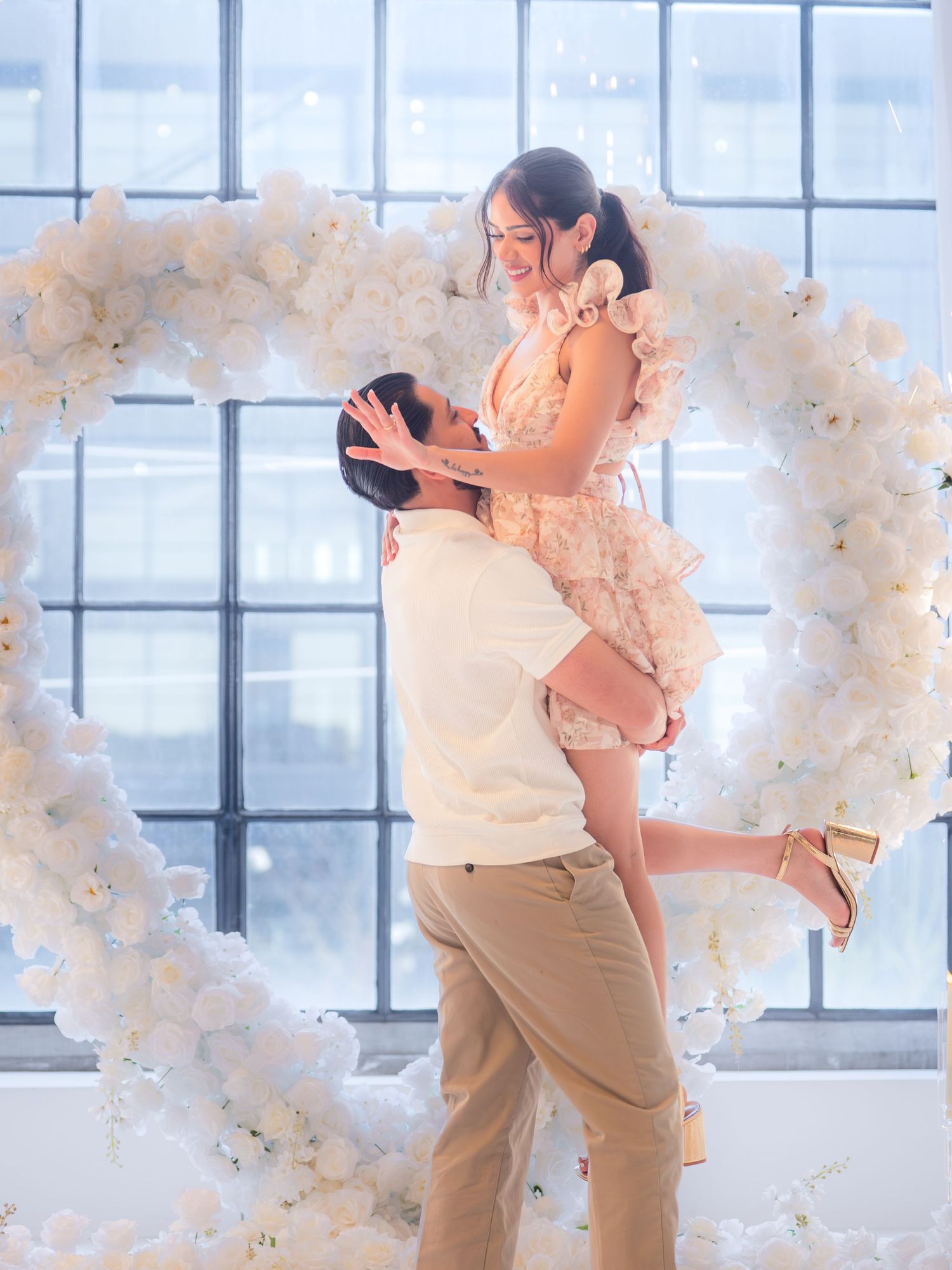 Couple celebrating engagement in front of white floral heart arch, man lifting woman in floral dress, romantic indoor setting.
