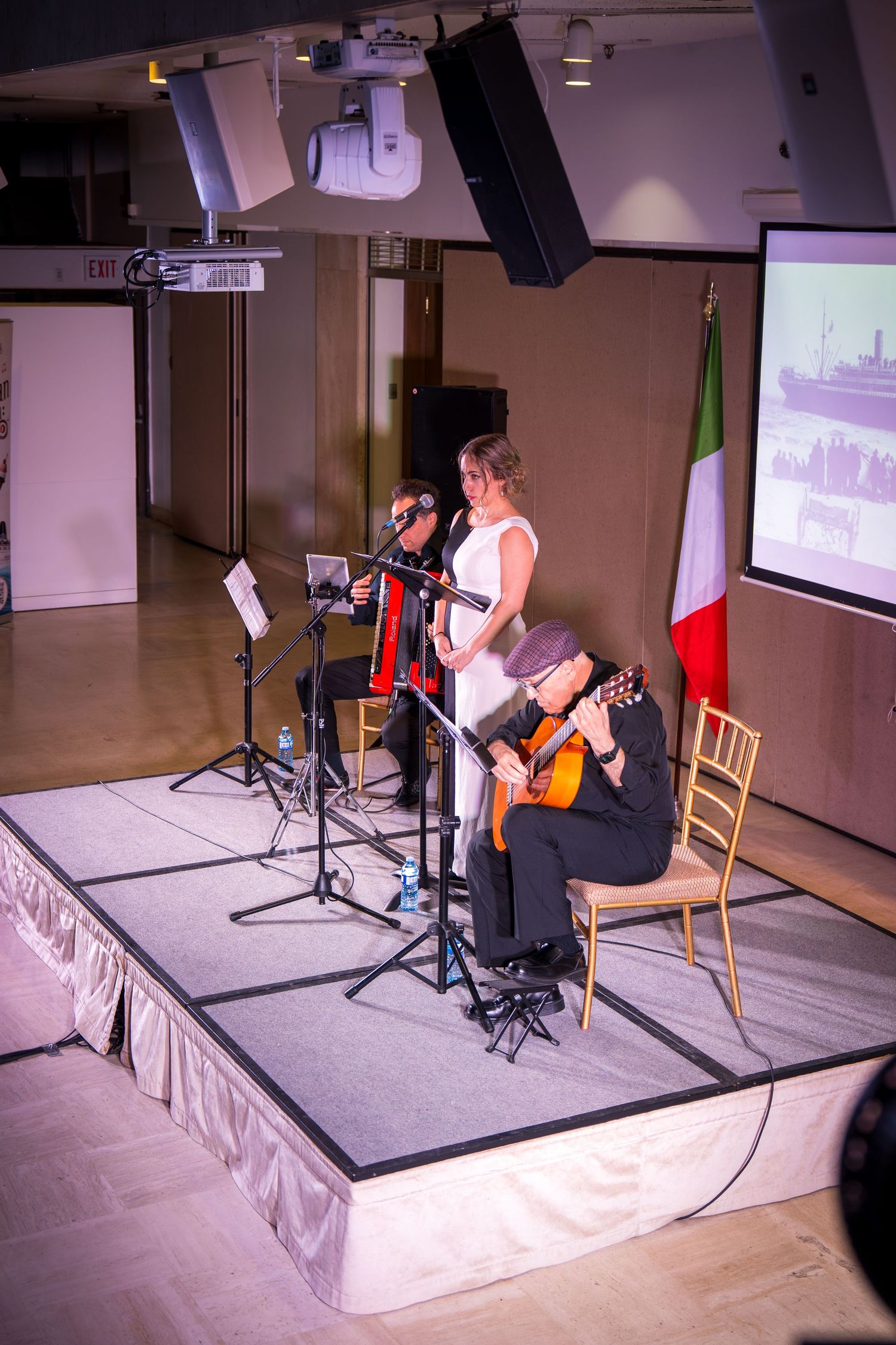 Three musicians perform on stage with guitar, accordion, and female vocalist beside an Italian flag.