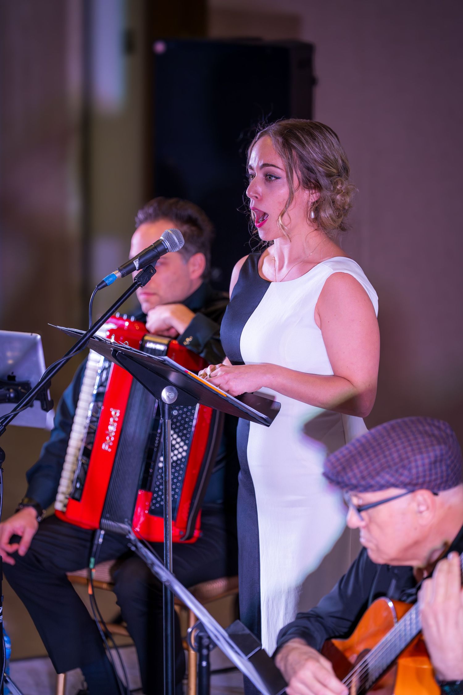 Female vocalist sings into microphone flanked by accordion and guitar players at a live performance.