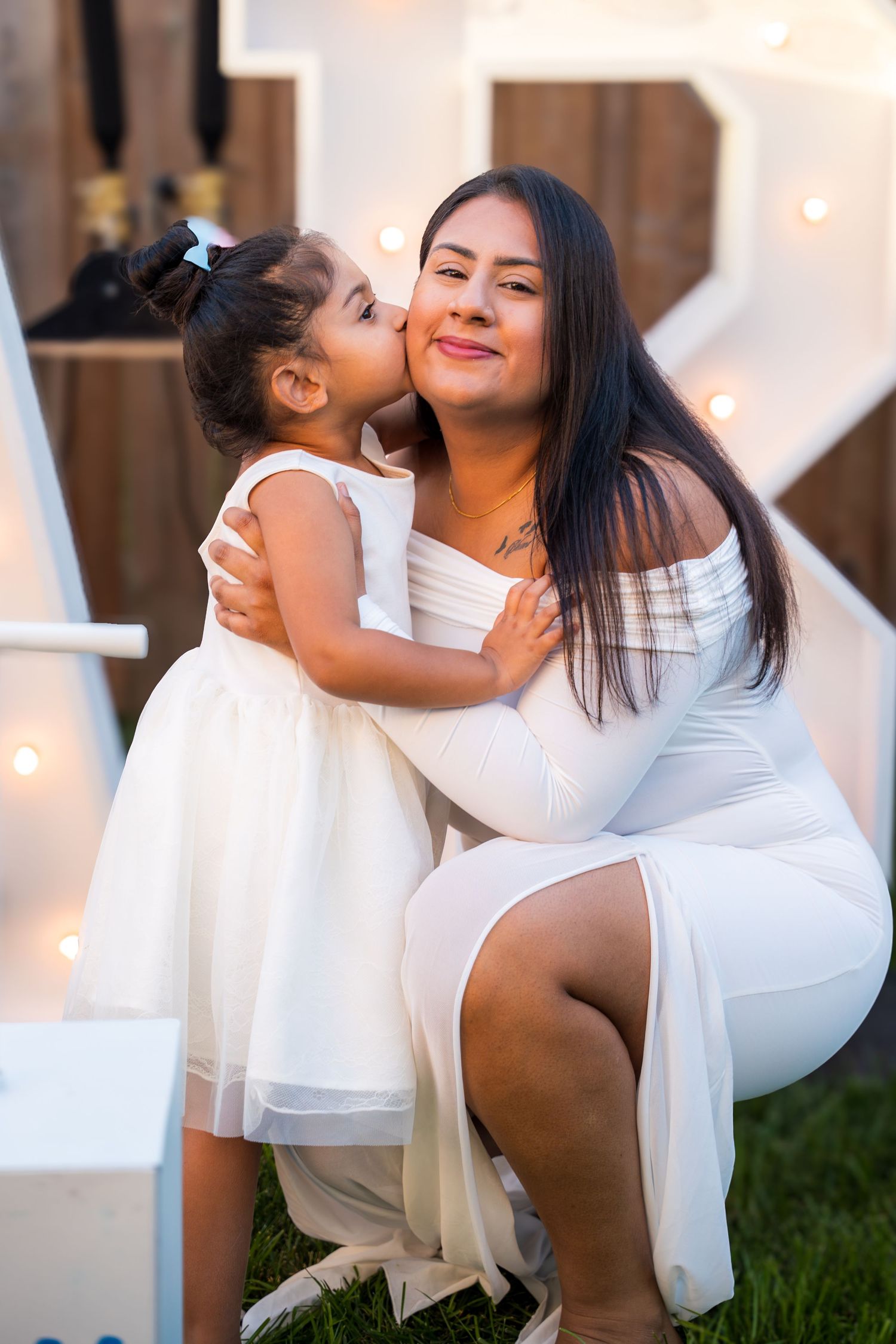 A mother and young daughter in matching white dresses share a sweet kiss at an outdoor celebration.