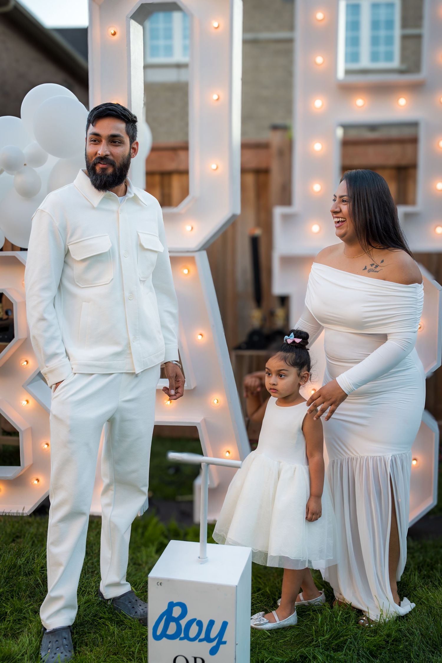 Family poses in white outfits at a boy gender reveal party with illuminated marquee letters.