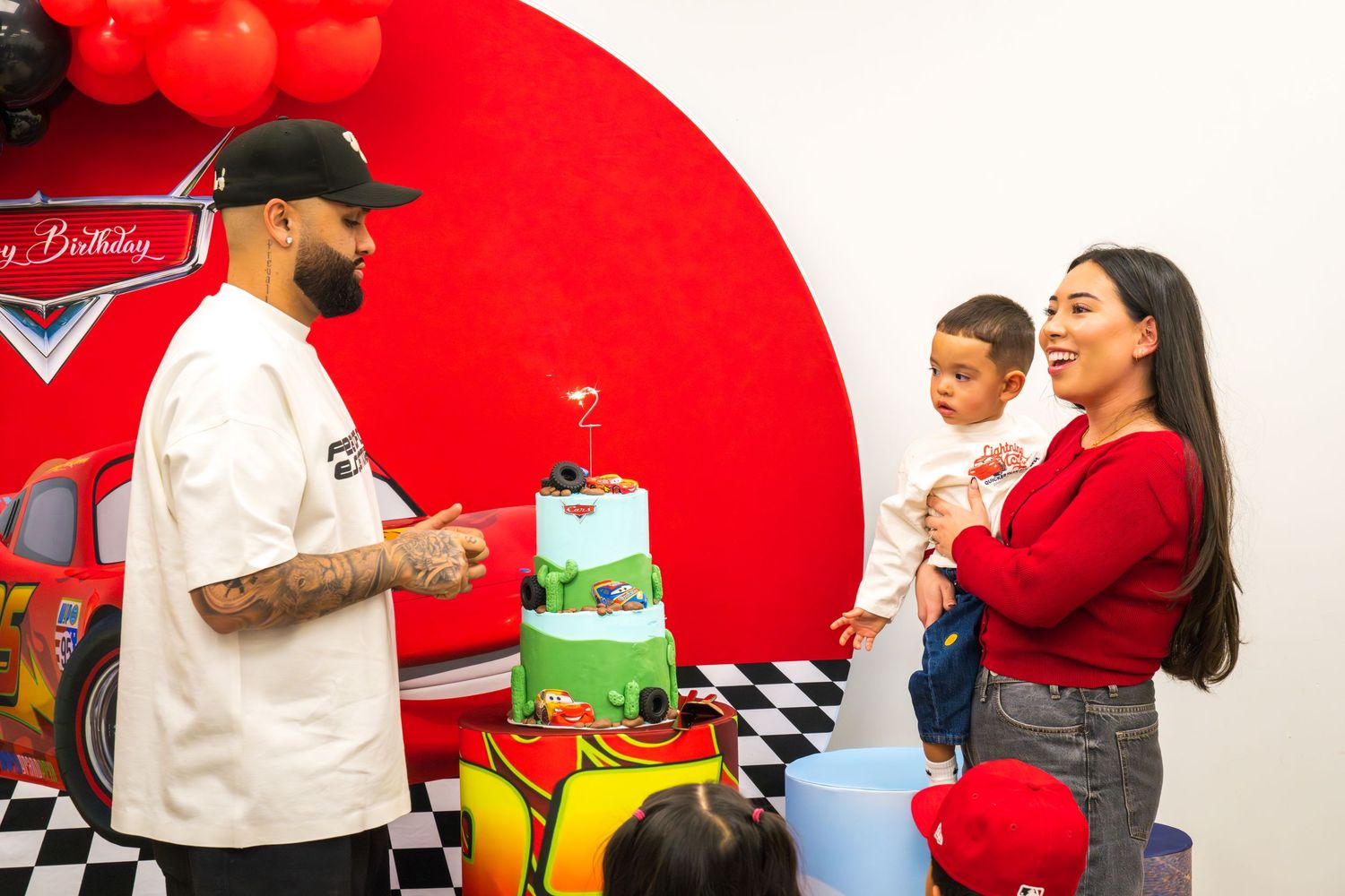 A man and woman with their baby celebrate in front of a Cars-themed birthday cake and decor.