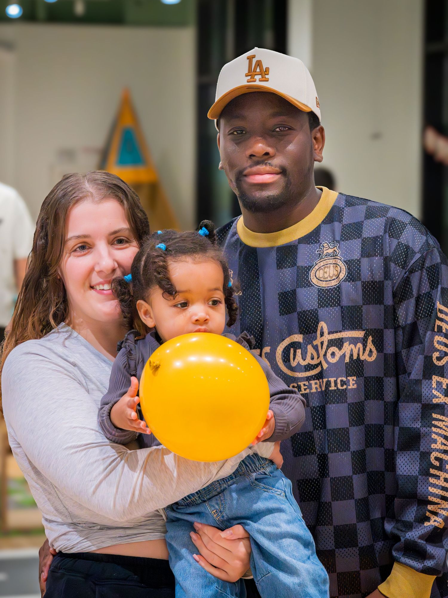 Smiling family with mother, father in LA cap, and toddler daughter holding a yellow balloon at a party.