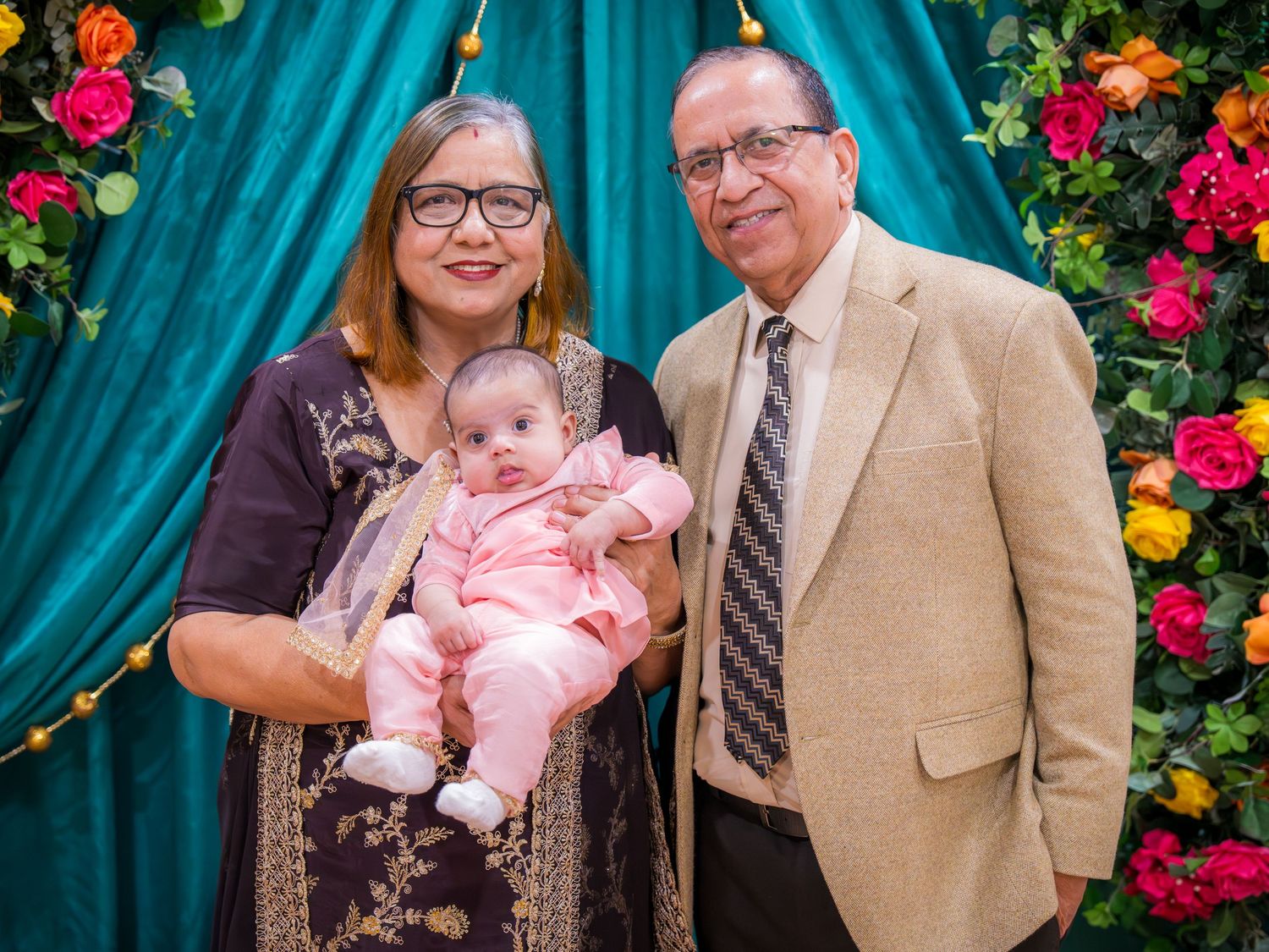 Older South Asian couple in formal attire holds pink-dressed infant at vibrant floral teal backdrop.
