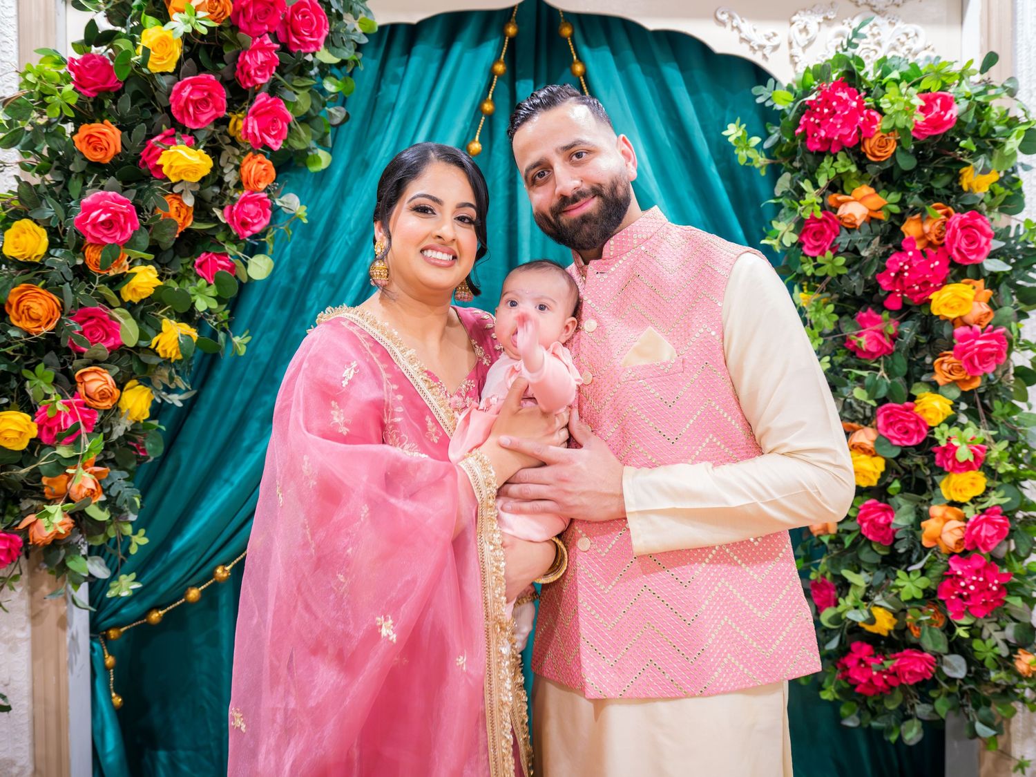 South Asian family in matching pink traditional attire with newborn at floral ceremony backdrop.