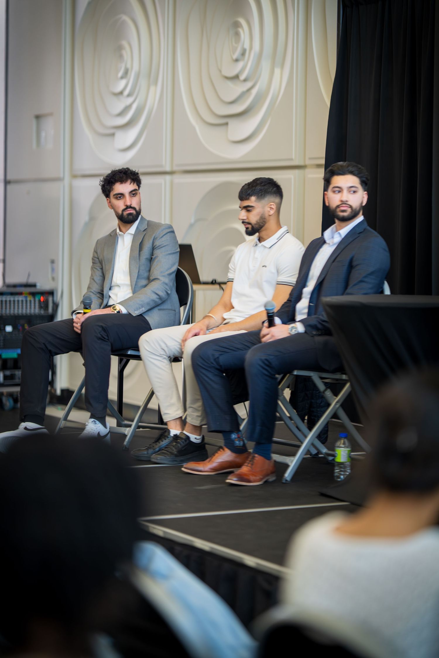 Three male panelists in business attire seated on folding chairs during professional event.