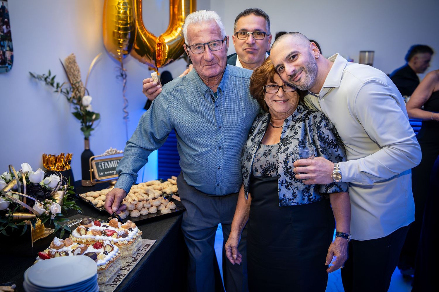 Three people pose together by a dessert table with gold balloon decorations visible.