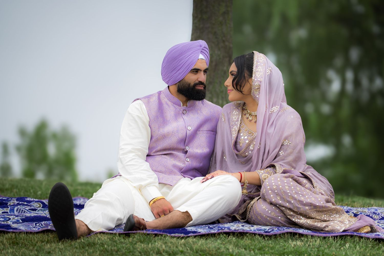 A couple in traditional Punjabi attire relaxing on a blanket outdoors surrounded by greenery.