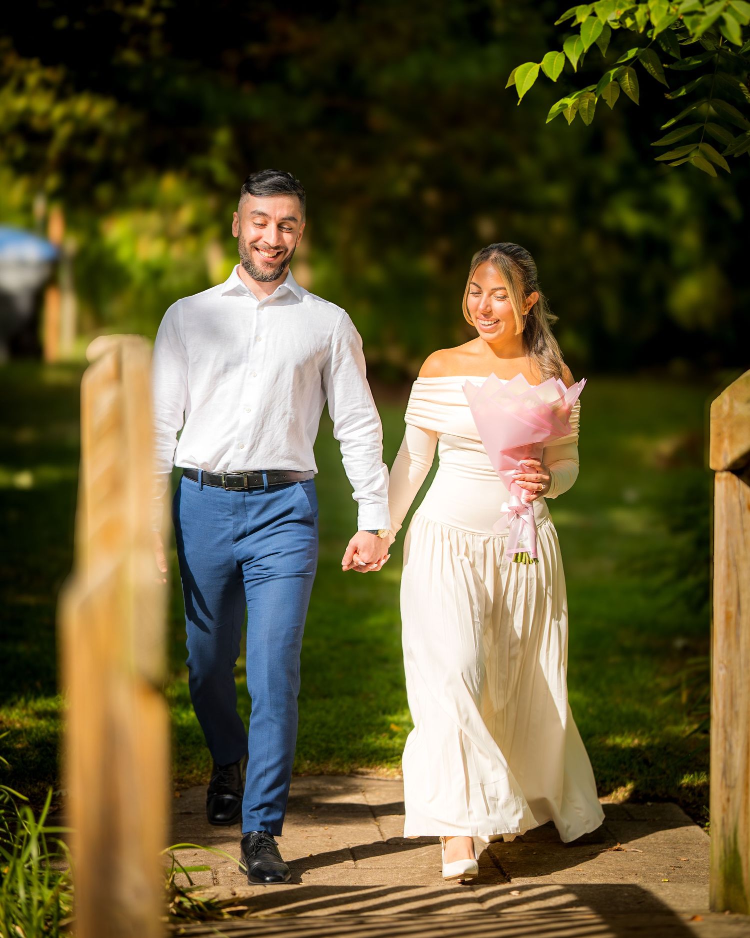 Couple walks hand in hand along a wooden path surrounded by greenery.
