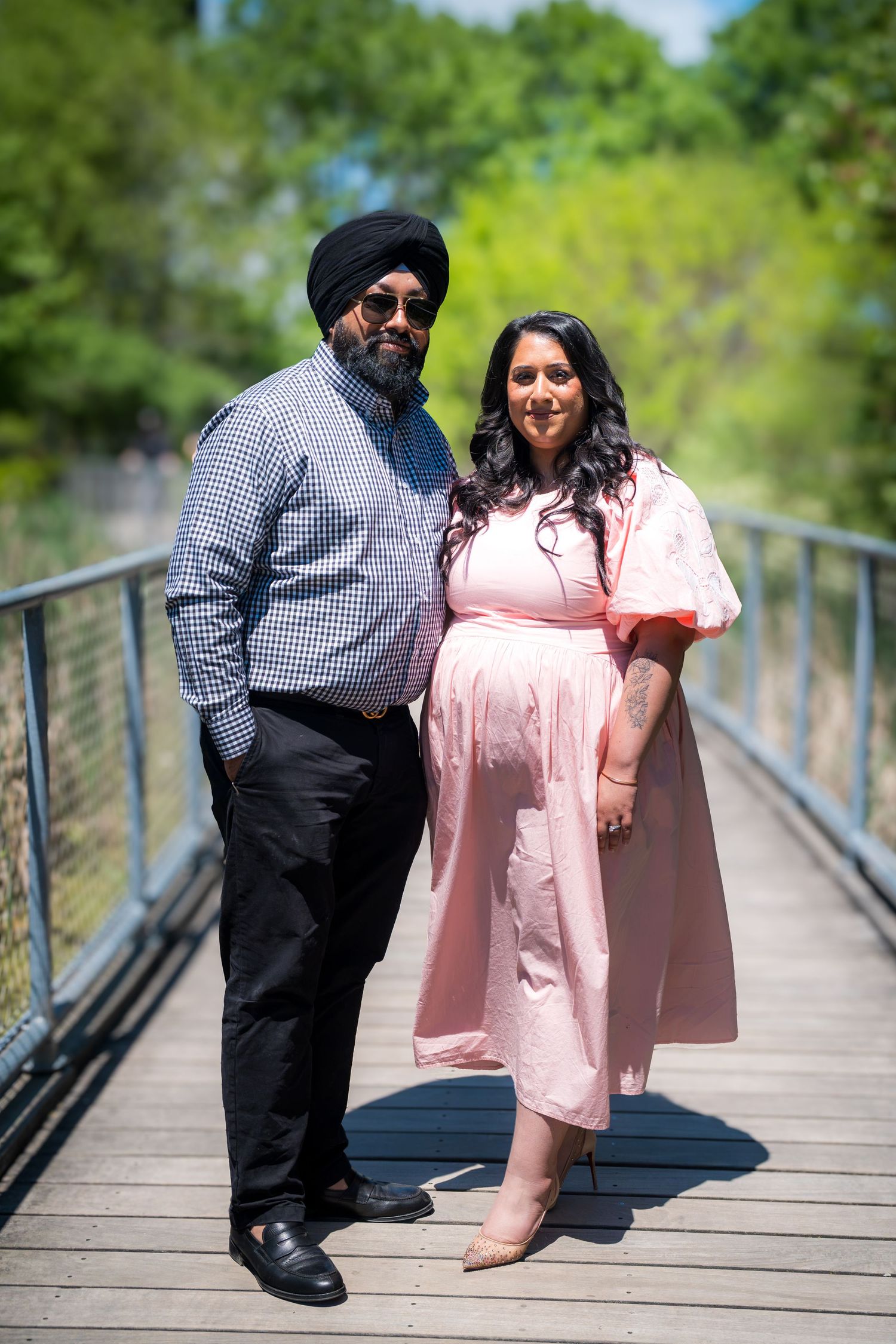 A pregnant woman in a pink dress poses with a man in a turban on a sunny wooden bridge surrounded by green trees.