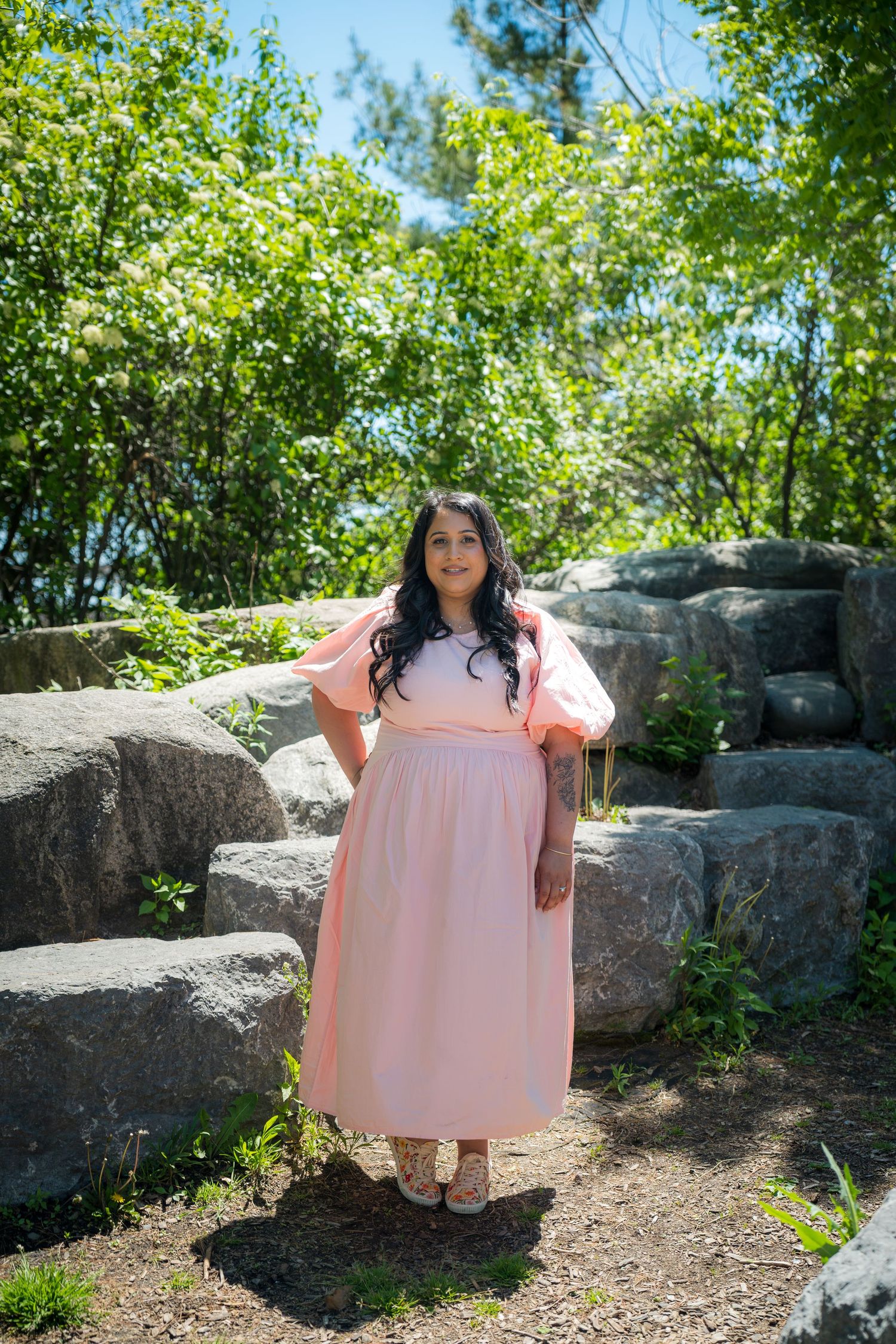 Pregnant woman in pink maxi dress with puff sleeves poses among rocks and greenery outdoors.