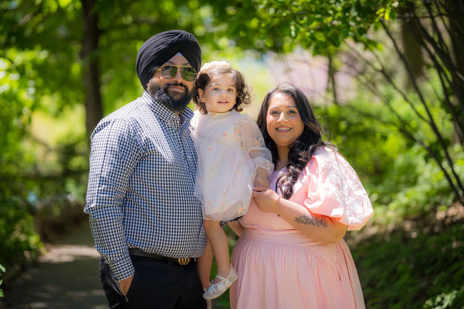 Sikh family with man in turban, woman in pink maternity dress, and toddler in white dress outdoors.