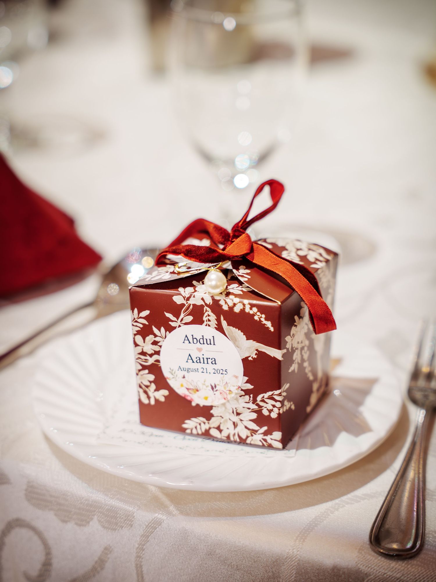 Red and white wedding favor boxes with floral designs displayed on white place settings.
