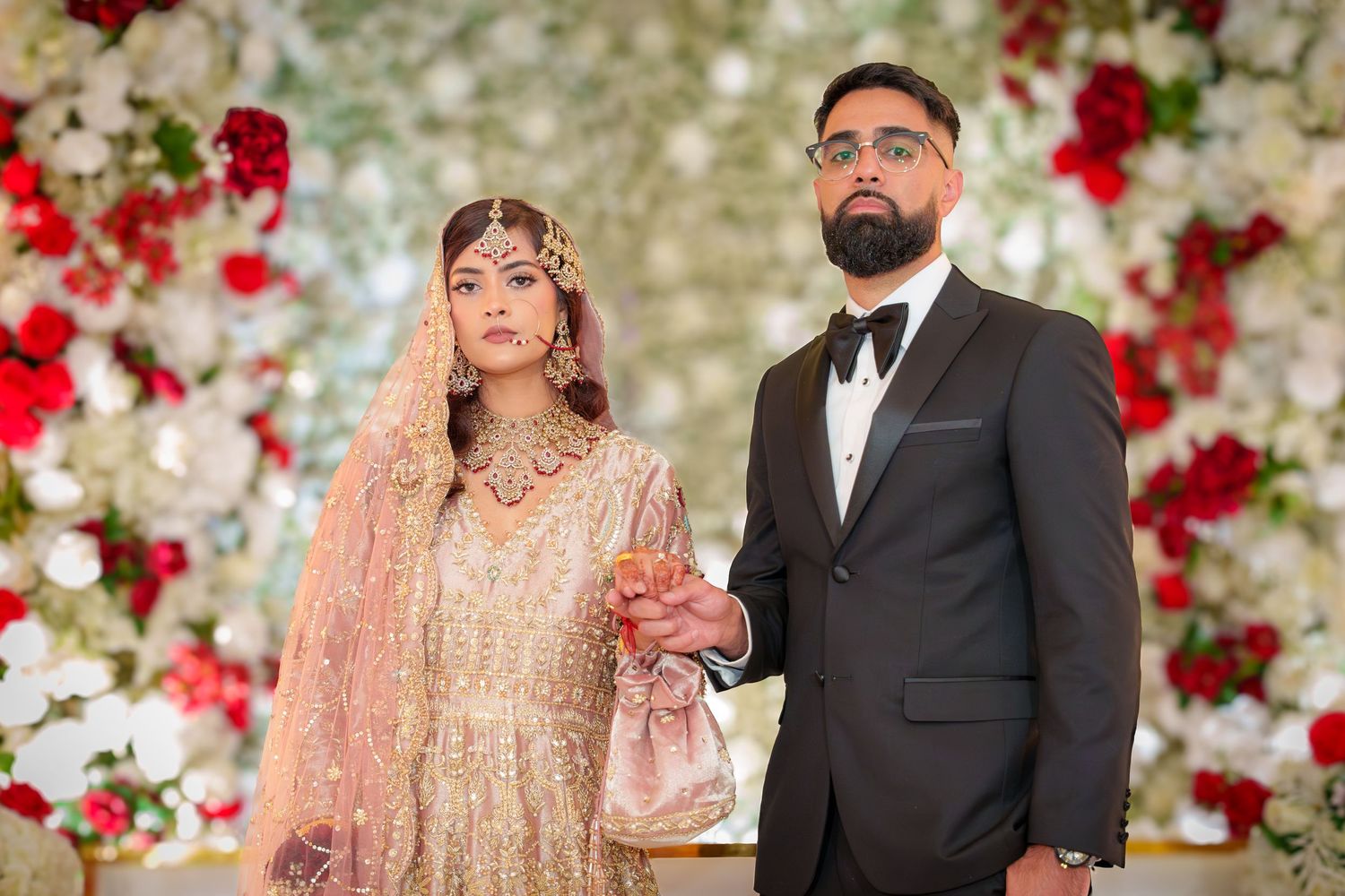 Wedding couple poses in front of luxurious floral wall with red roses.