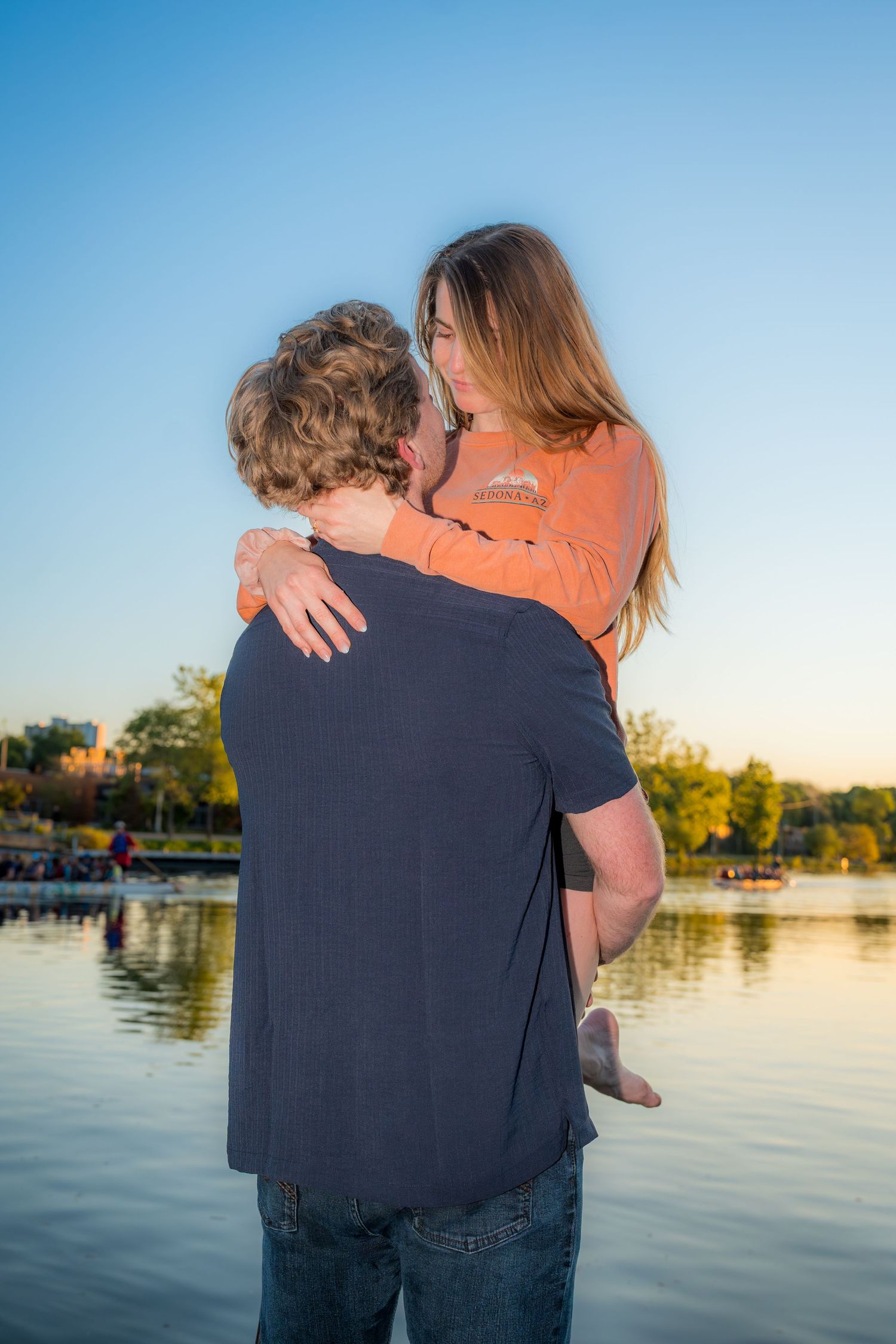 Man lifting woman in his arms near a lake at golden hour, sharing a tender embrace.