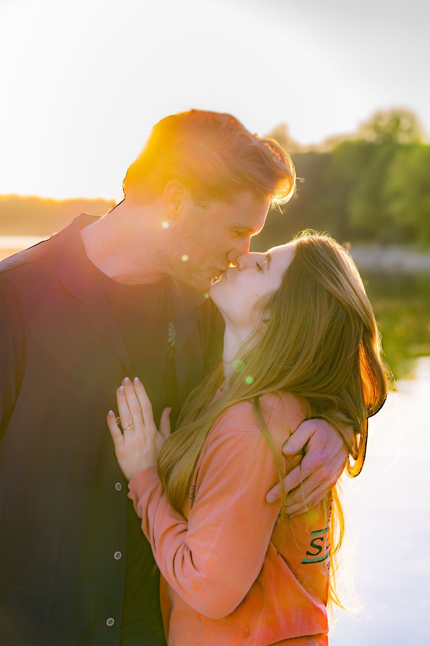 Romantic couple sharing a kiss at golden sunset by a lake, woman in coral top embraced by man in dark shirt.