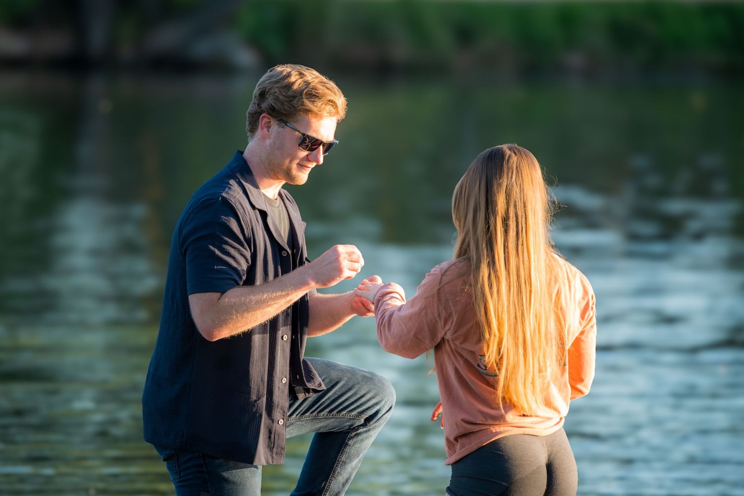 Man kneeling by a lake proposing to a woman in a pink hoodie with a ring outdoors.