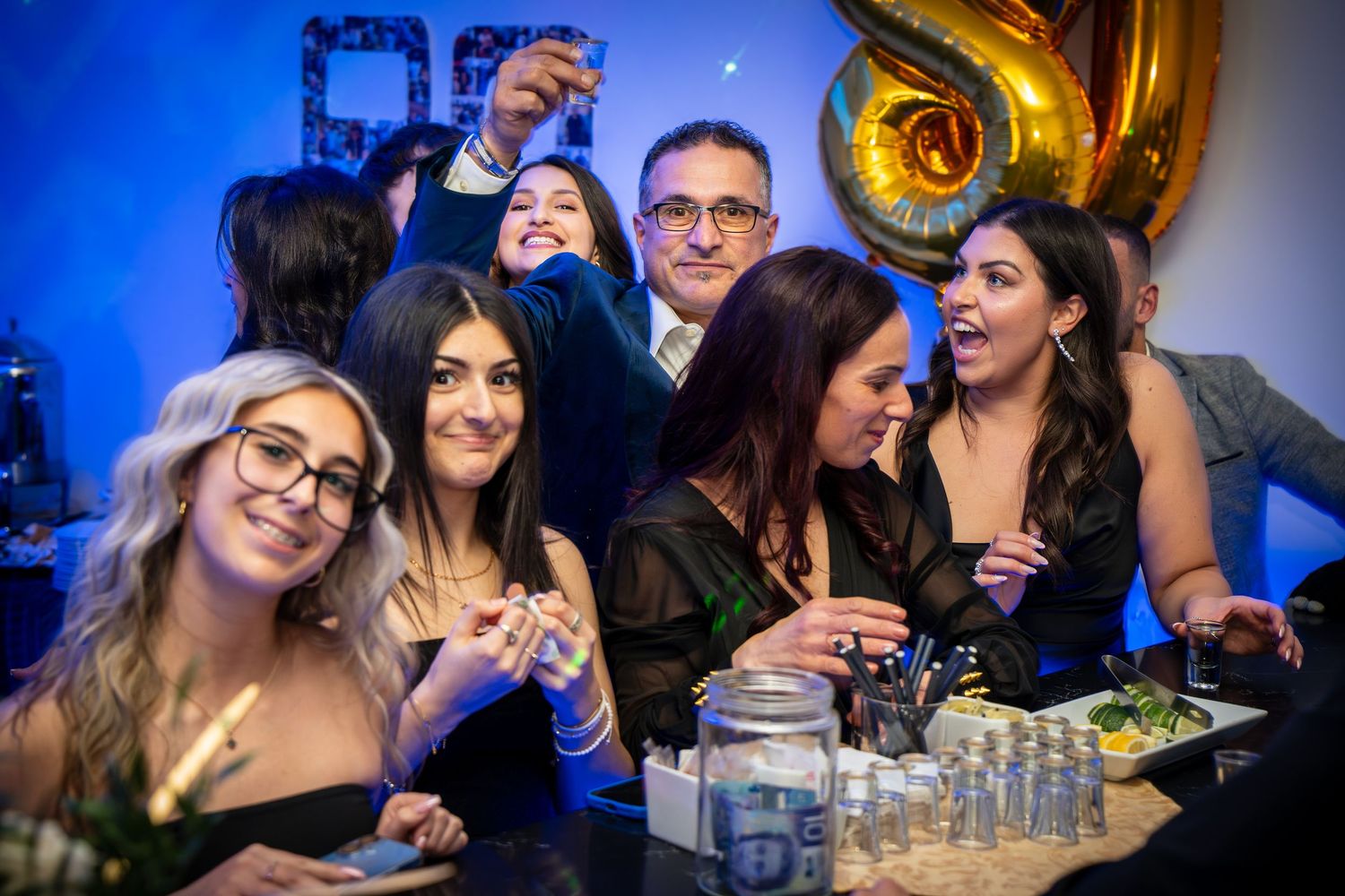 Friends gathered around a table celebrating with drinks under blue mood lighting and gold balloons.