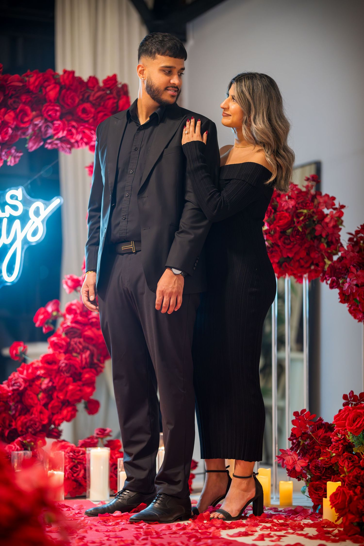 Elegant couple in formal black outfits stand together surrounded by bright red floral arrangements.