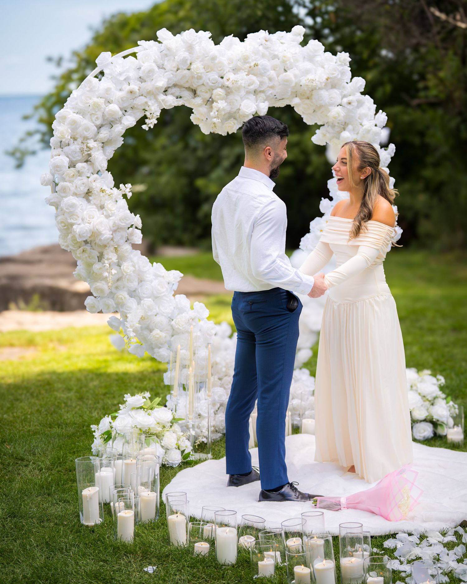 Wedding ceremony setup with white floral arch and glass candle holders on grass.
