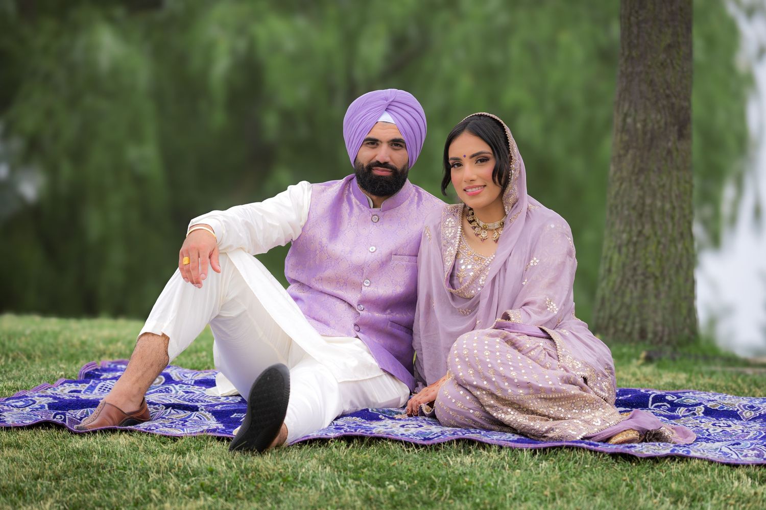 Three people relax outdoors on a patterned blue blanket wearing coordinated traditional Punjabi attire.