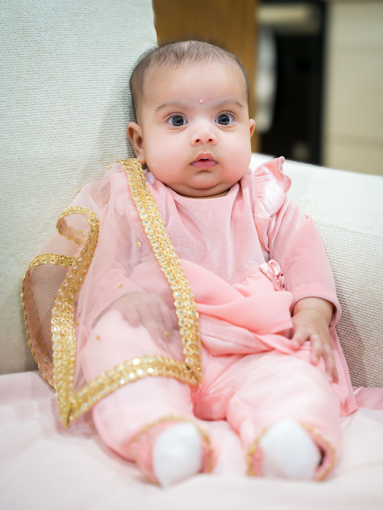 Adorable baby girl in pink traditional Indian outfit with gold trim dupatta sitting upright.