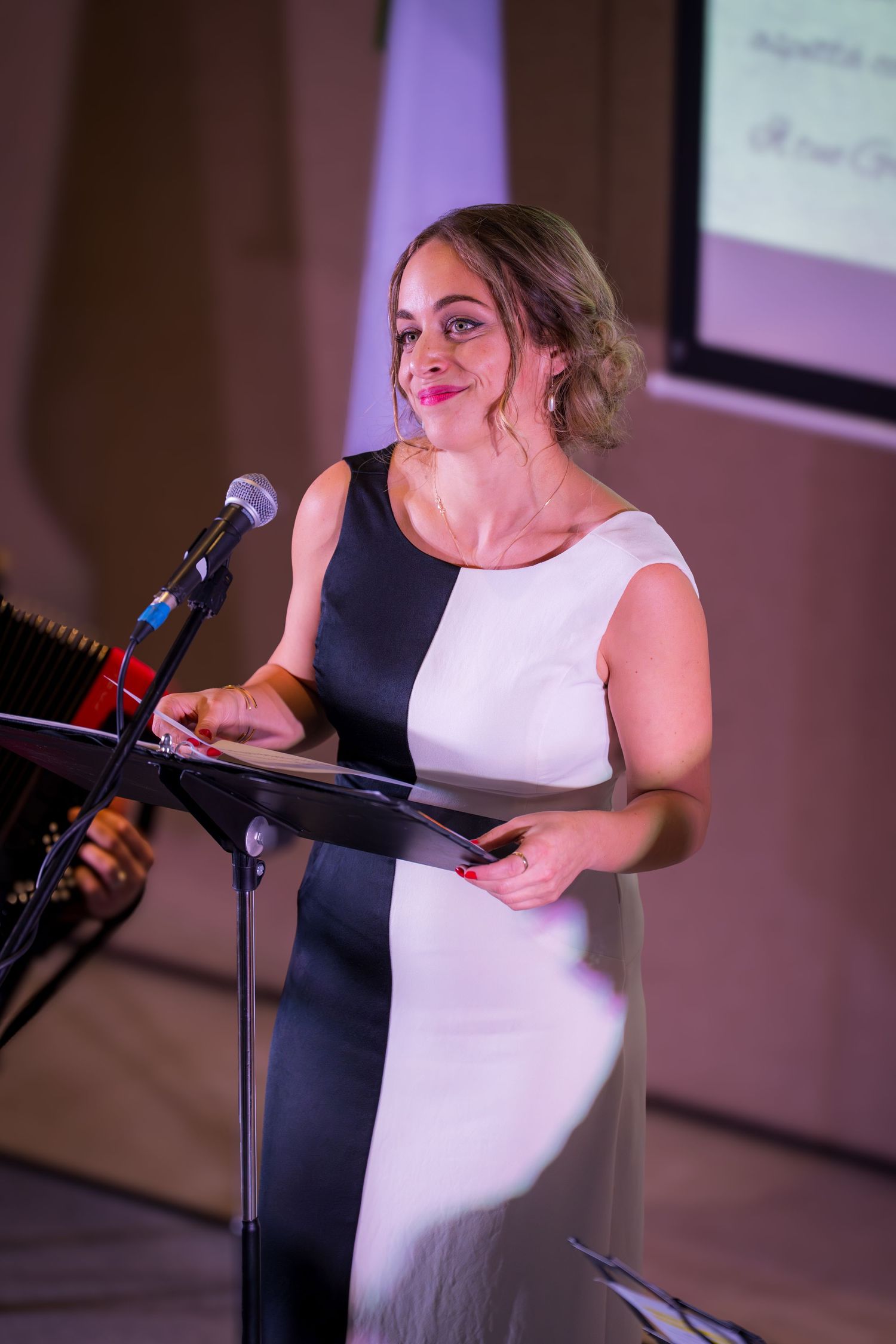 Smiling woman in black and white dress speaking at a podium microphone during a live event.