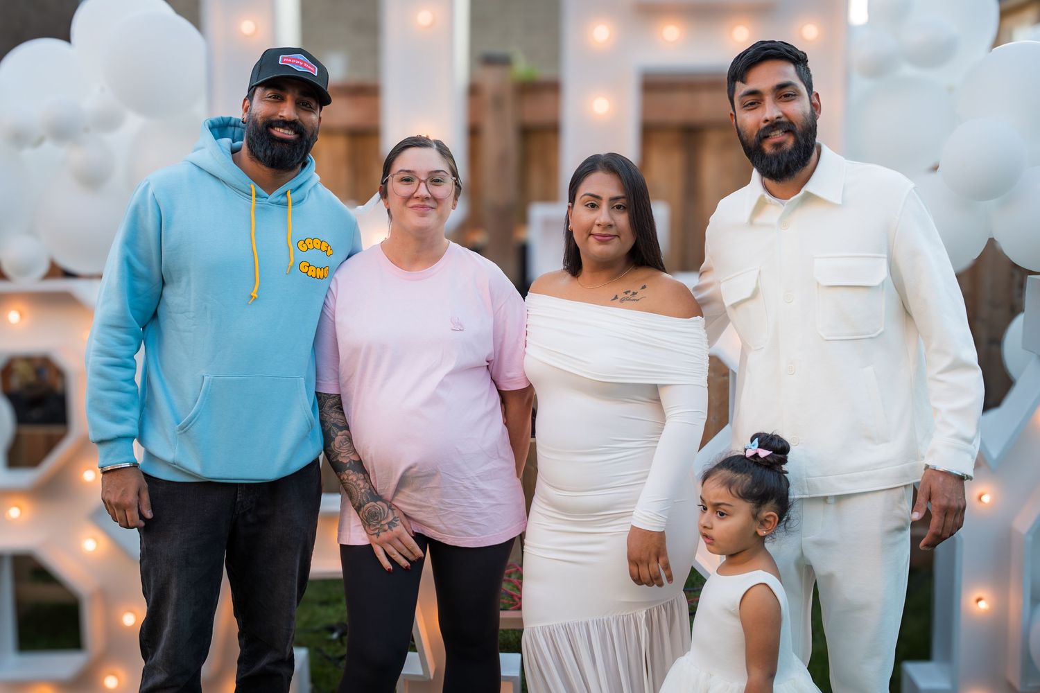 Group of five adults and child pose together at outdoor baby shower with white balloon decorations.