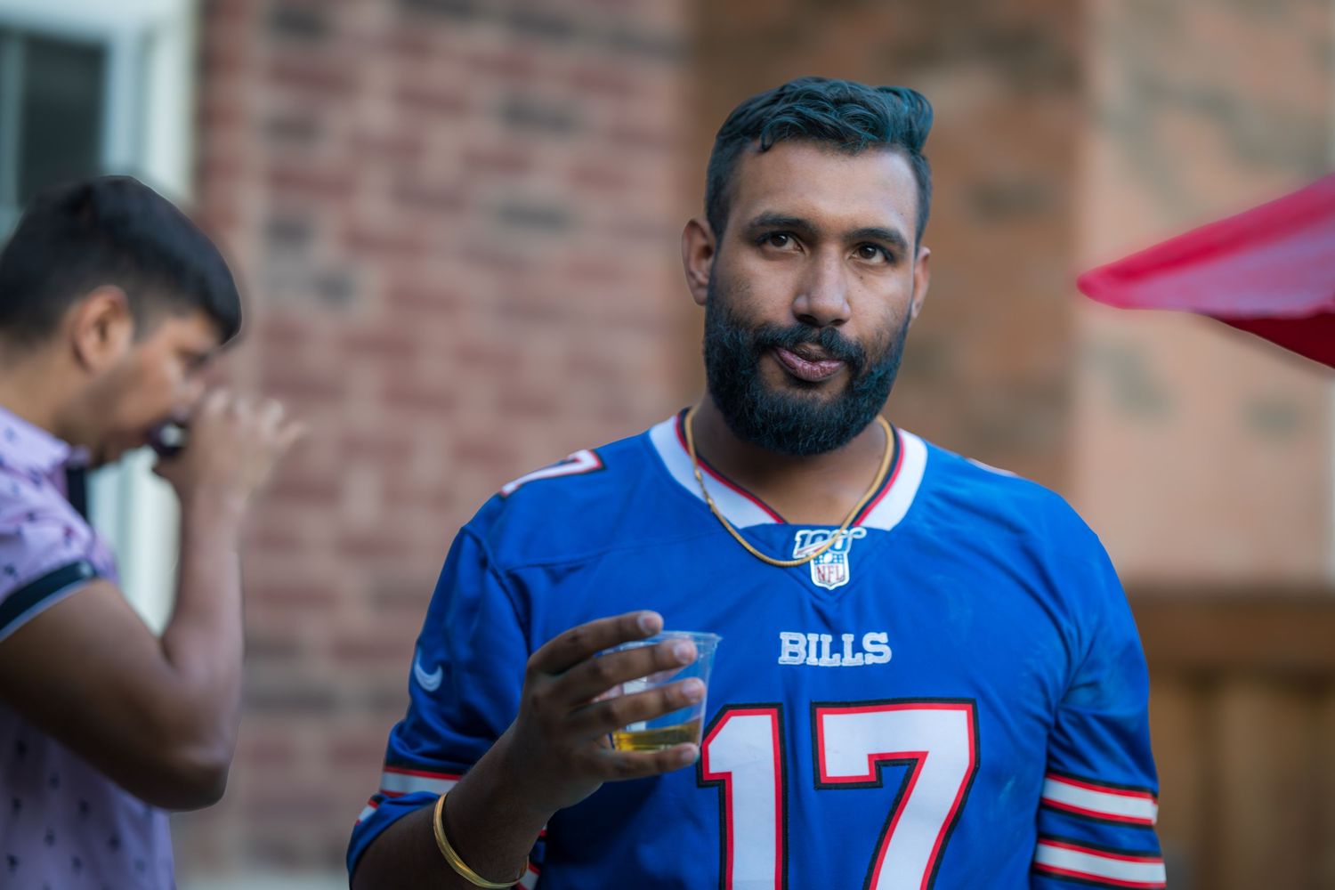 A bearded man wearing a Buffalo Bills number 17 jersey holds a drink at an outdoor backyard gathering.