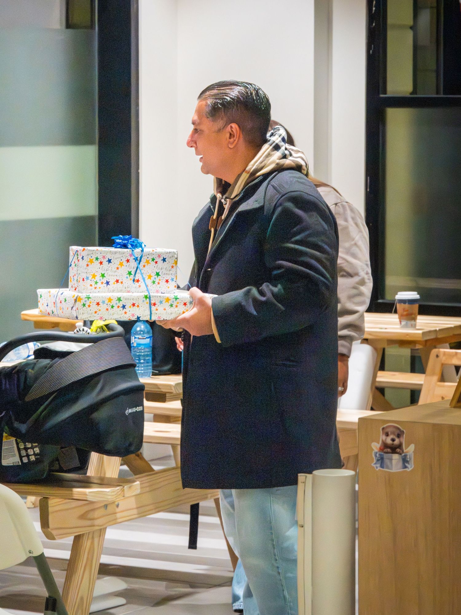 A man in a navy coat carries a colorful polka-dot gift box at an indoor birthday celebration.