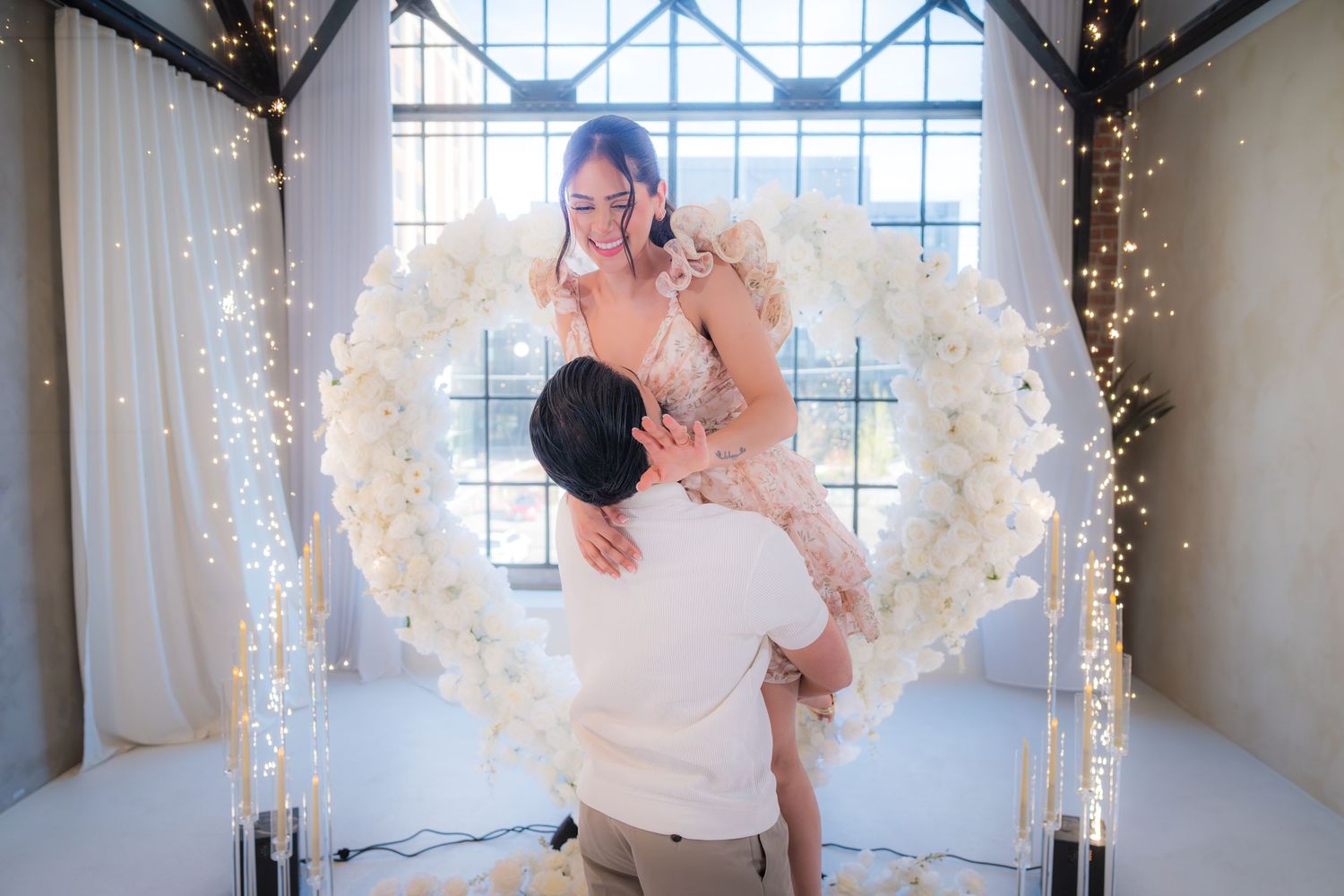 A man lifts his overjoyed fiancee in front of a stunning white floral heart arch with fairy lights.