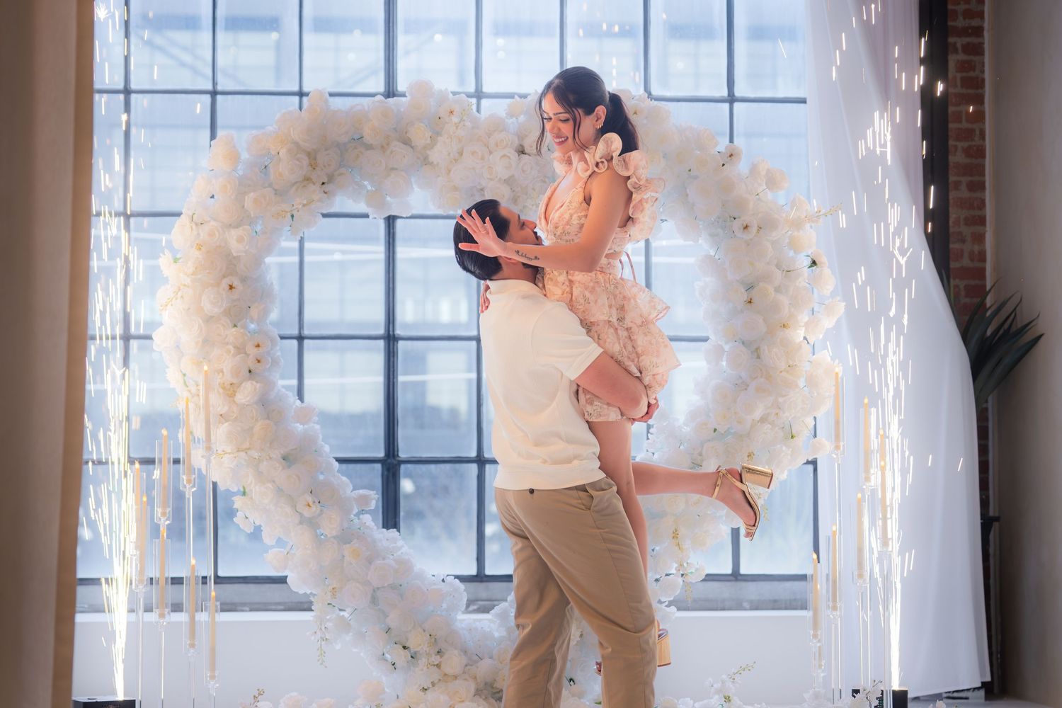Man lifting woman in front of a white floral heart arch with sparkling fireworks backdrop.