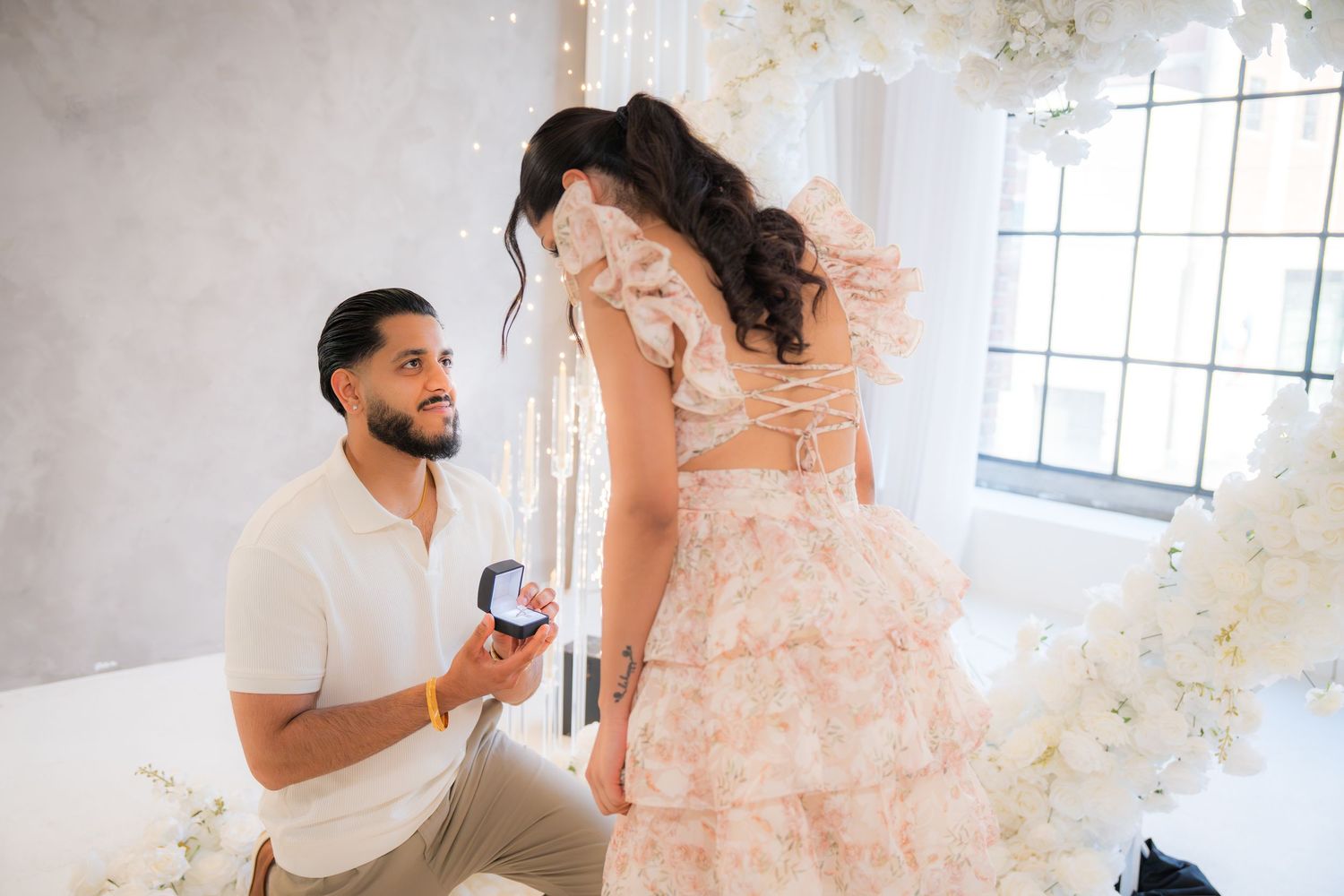 Man proposing on one knee holding ring box to woman in pink ruffled dress.