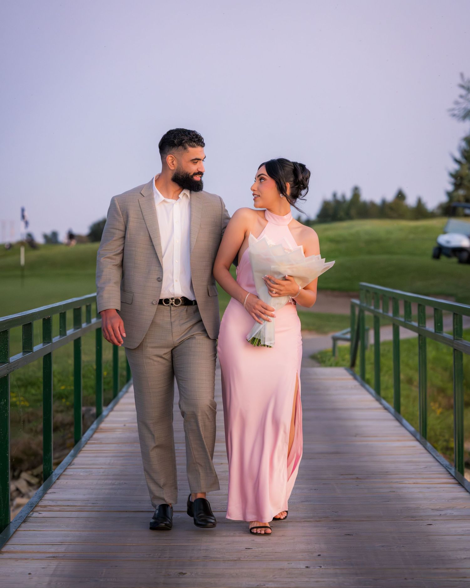 A couple in formal attire poses on a wooden bridge with green railings.