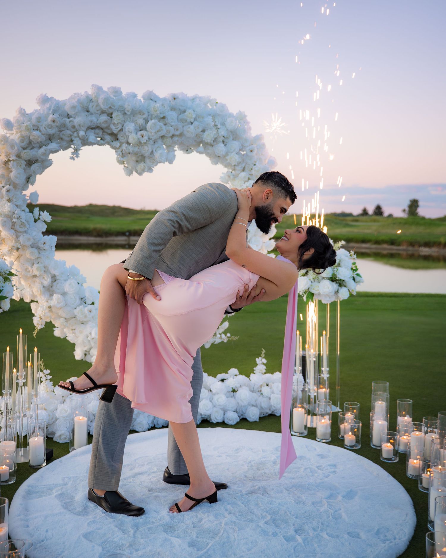 Romantic dip pose during sunset proposal under white floral arch surrounded by glowing candles.