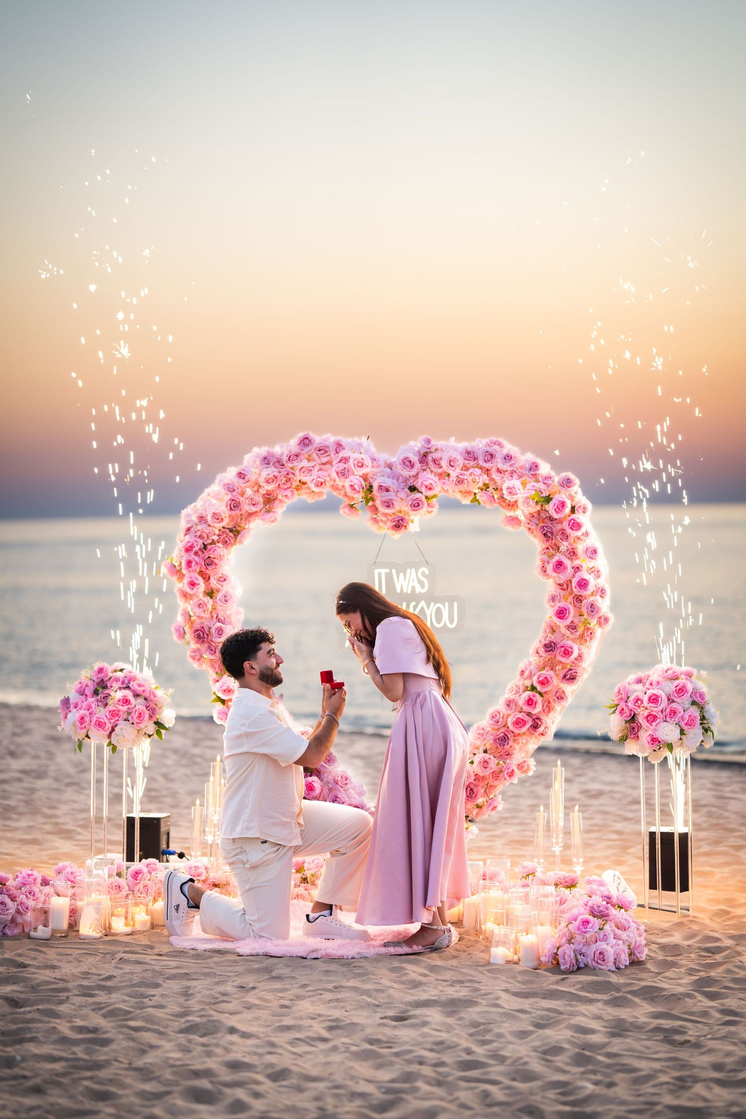 A magical proposal scene on the beach with sparkling lights and pink heart-shaped flower arch at sunset.