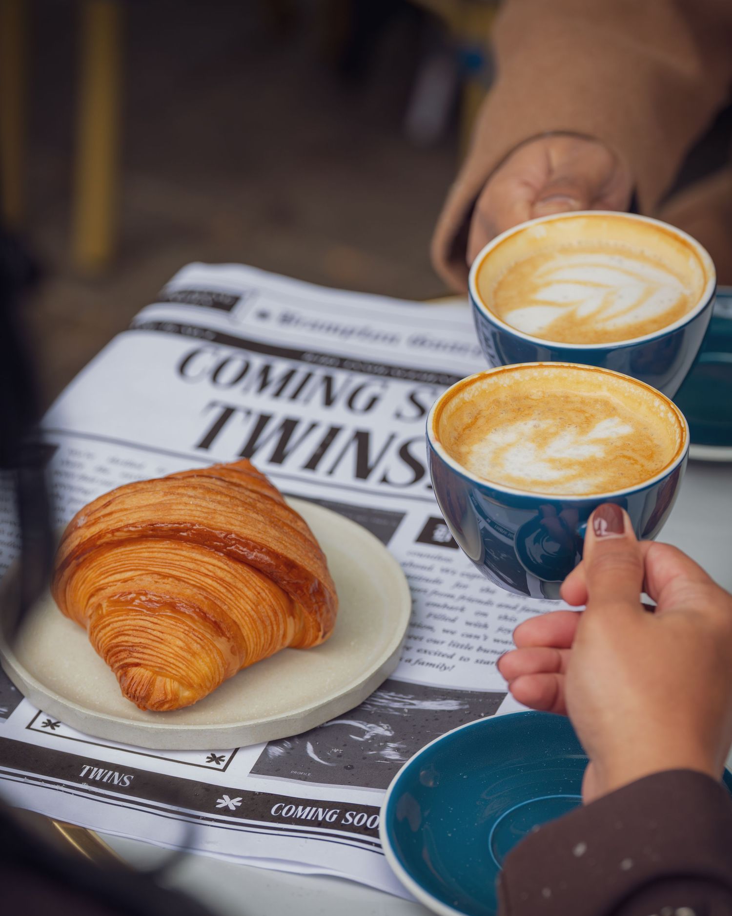 Coffee cups and croissants on plates next to a newspaper showing the word 'TWINS'.