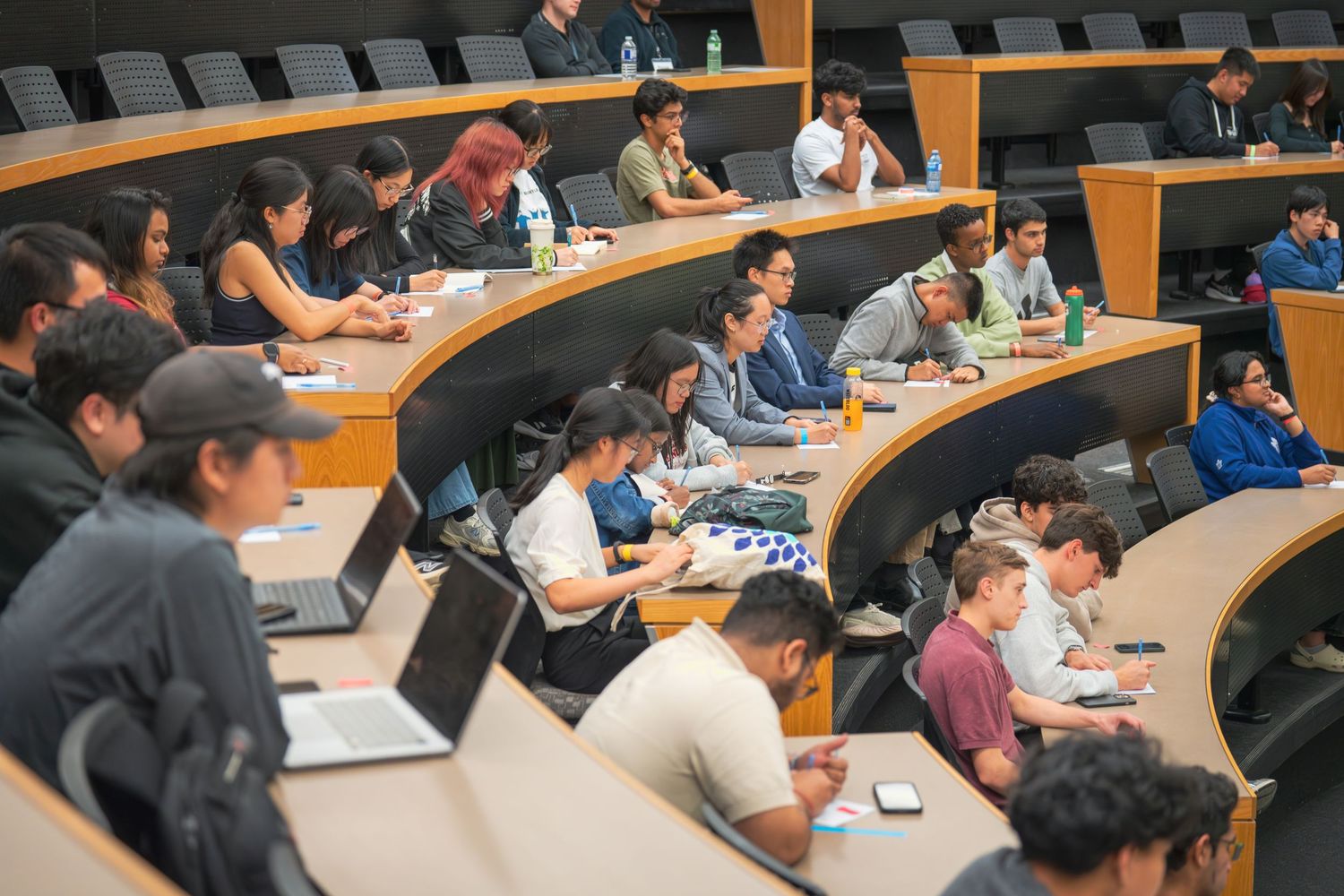 Students take notes in curved lecture hall seating during presentation.