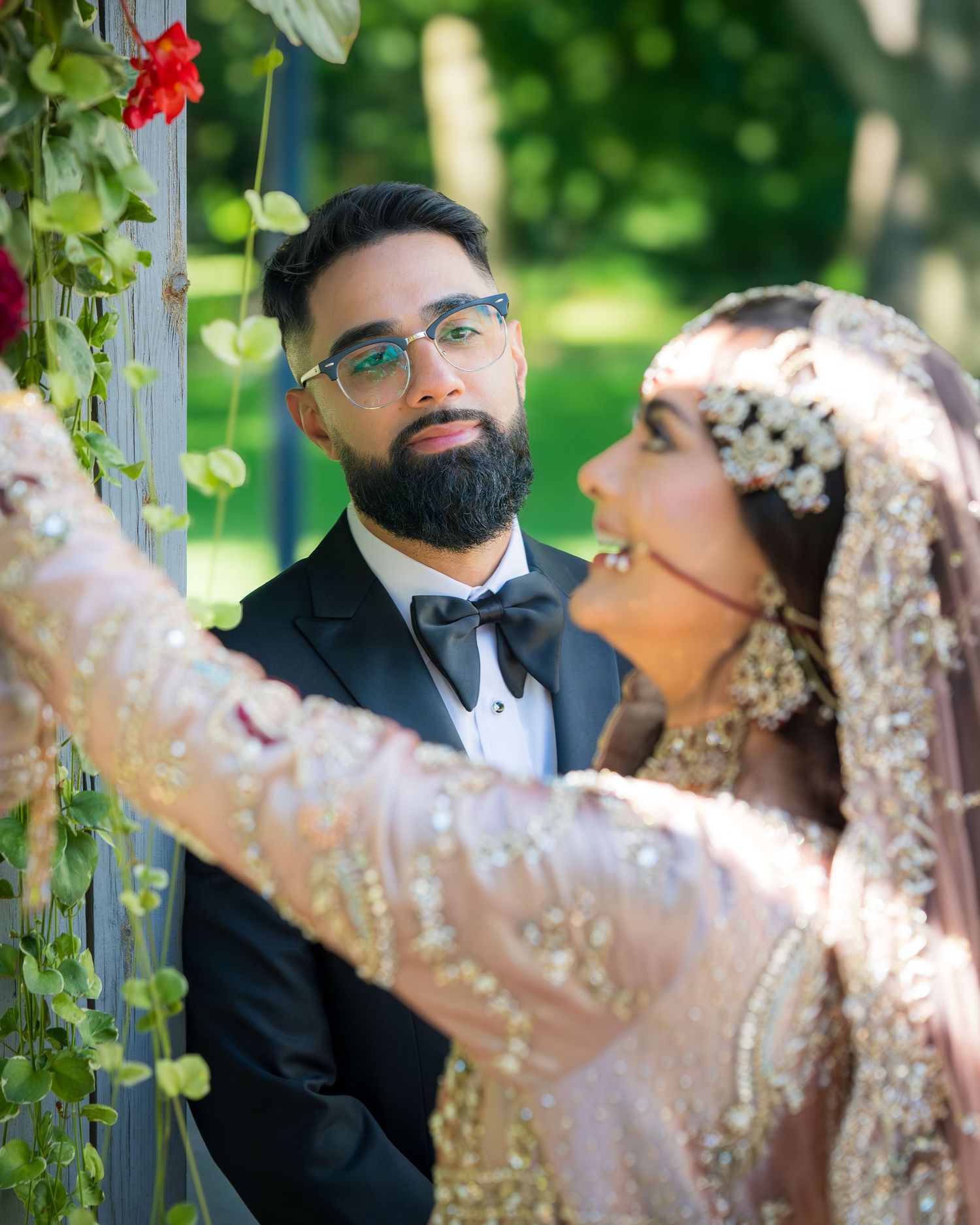 Traditional South Asian wedding couple poses outdoors with red flowers and greenery in the background.